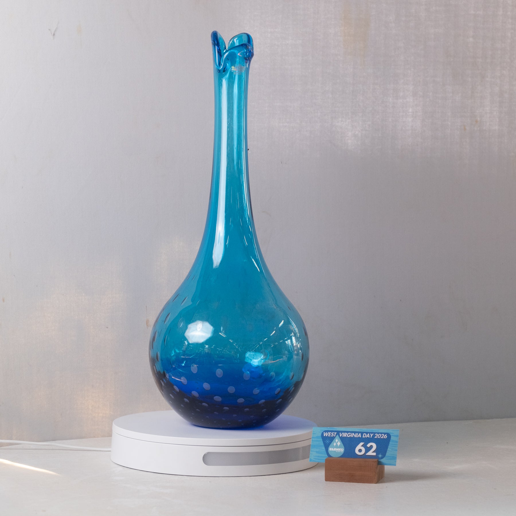 A blue glass vase with a flared neck and dotted pattern near the base, displayed on a white circular light box with a small blue price card in front, against a light gray fabric backdrop.