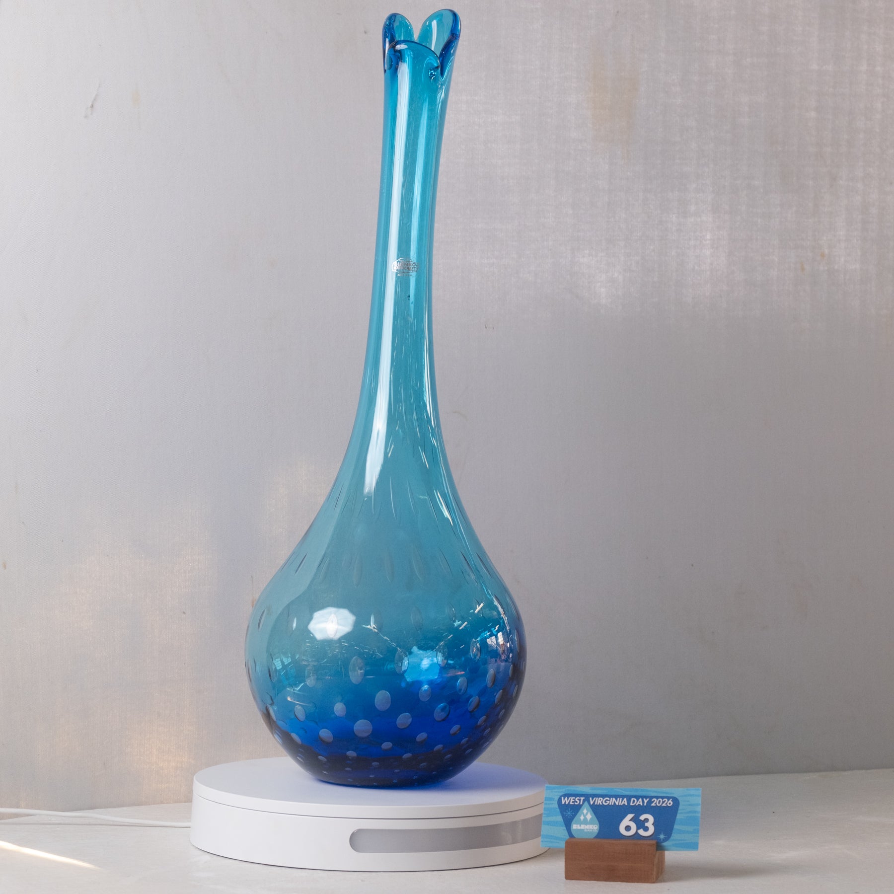 A blue glass vase with a long, flared neck and a rounded base, displayed on a white circular pedestal. The base of the vase has small white speckles, and a small blue and white size card is placed next to it on a wooden stand.