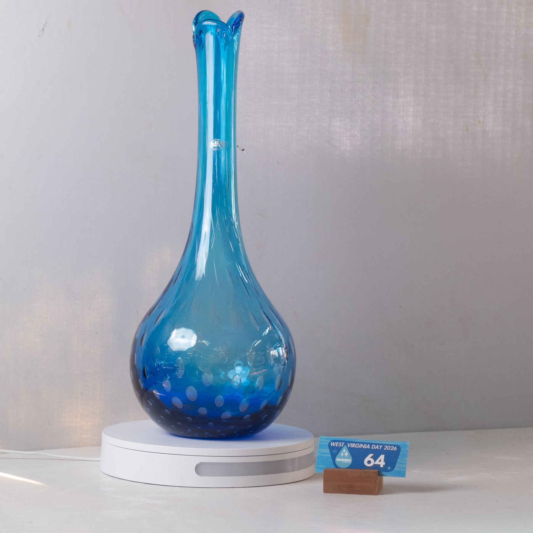 A translucent blue glass vase with a flared neck and rounded bulbous base, displayed on a white circular light box. A small blue inventory card is placed beside it on a white surface against a light gray backdrop.