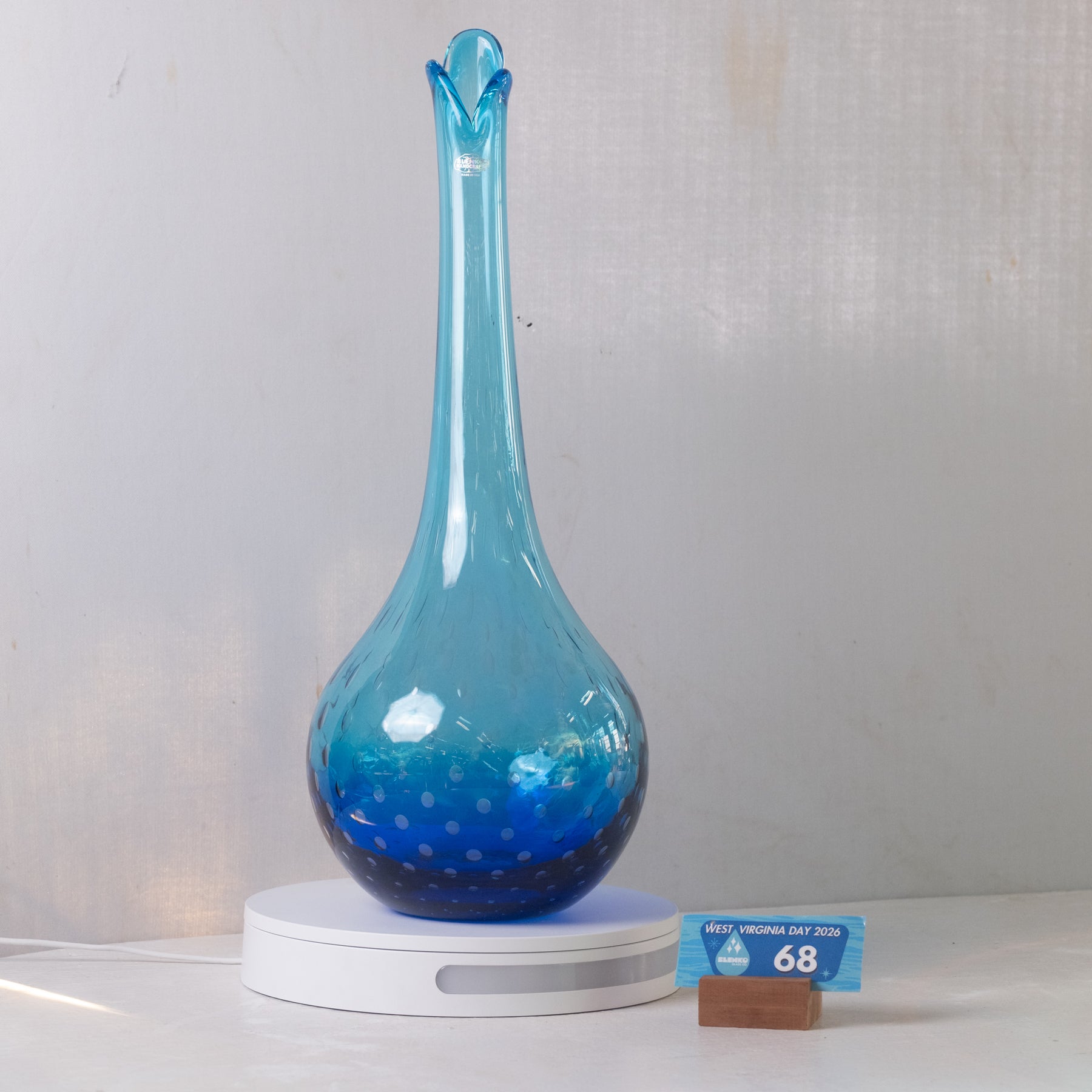 A blue glass diffuser vase with a long neck and a white circular base, shown on a light gray background with a small price tag in front.