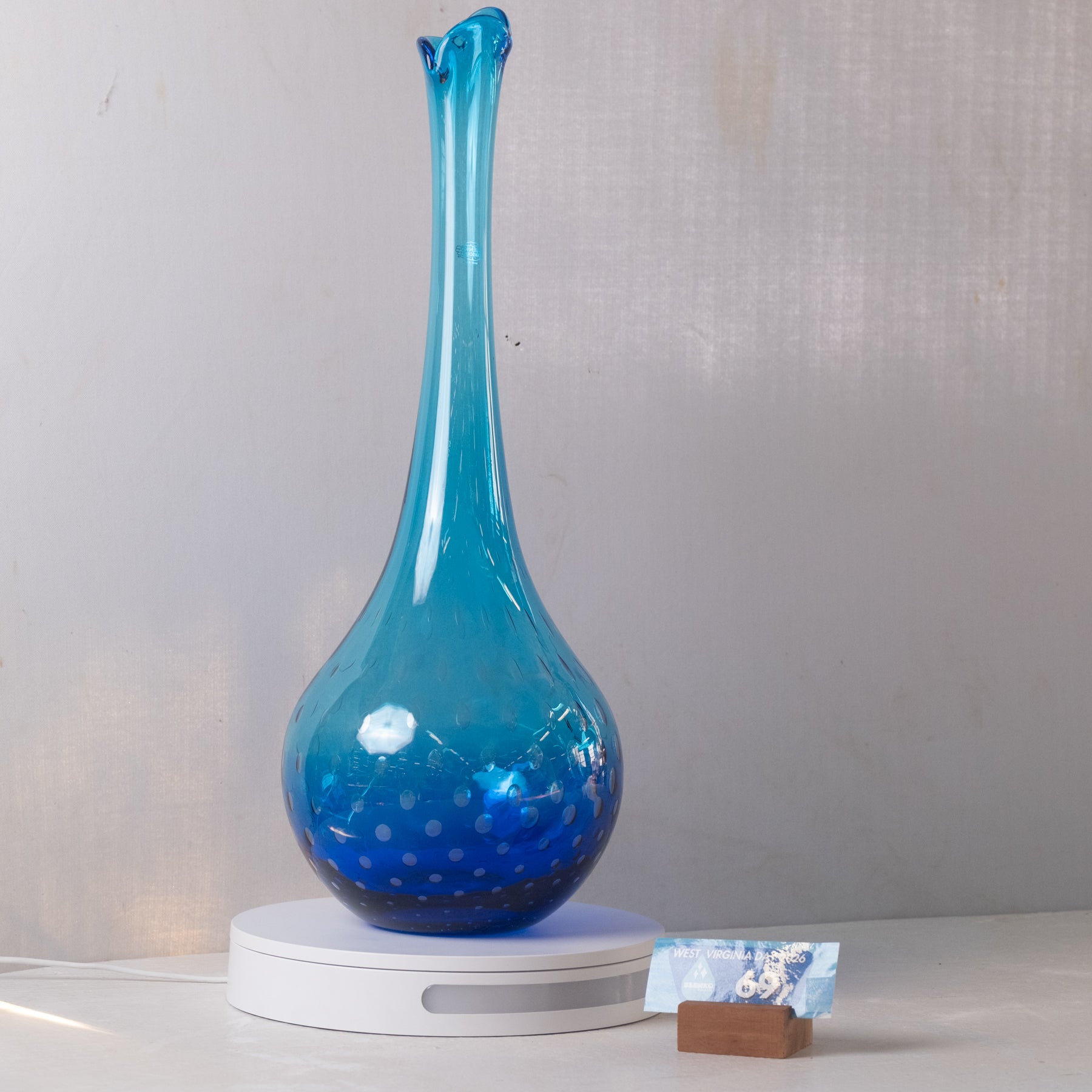 A blue glass vase with a flared neck and bubble pattern, displayed on a white circular base against a light gray wall.