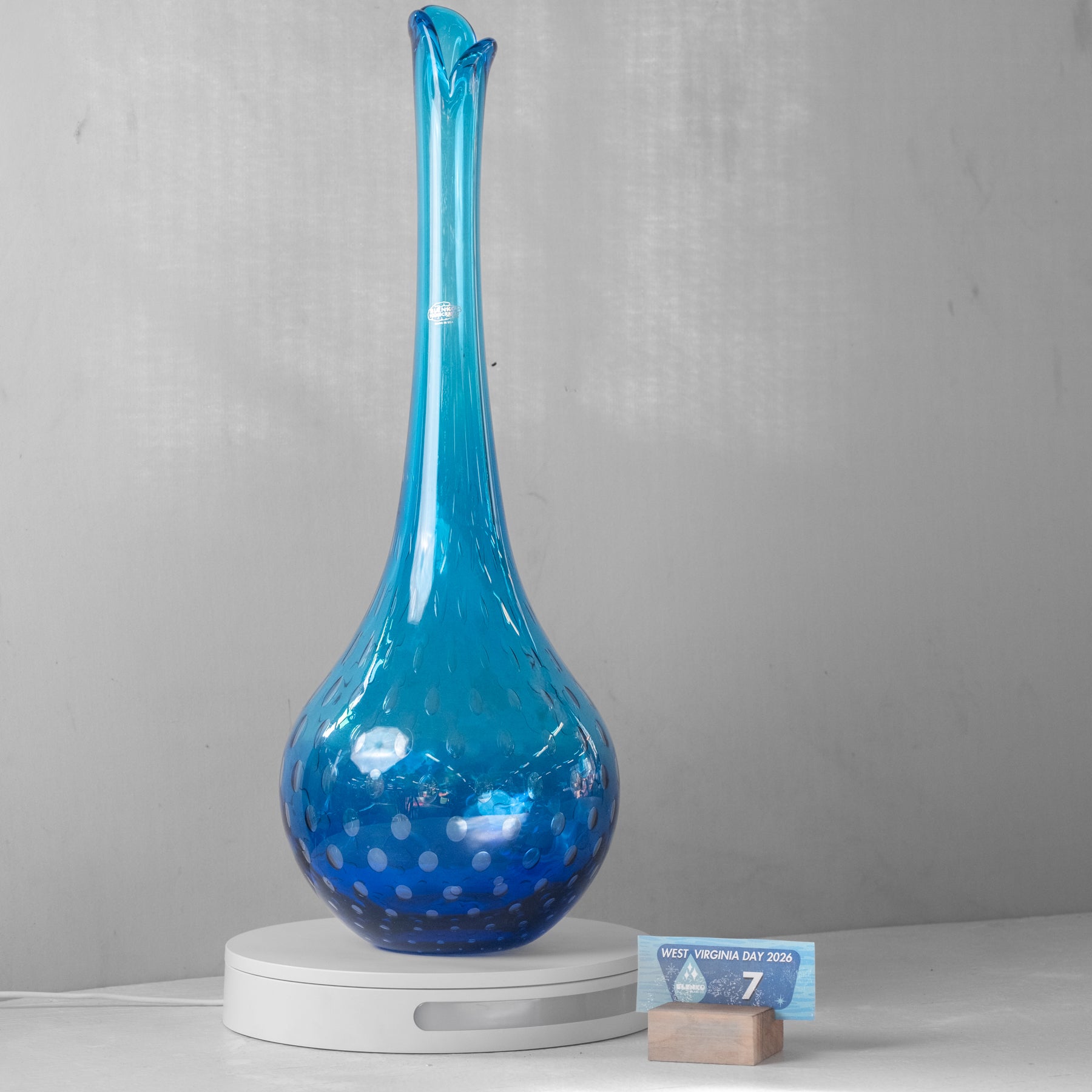 A tall, flared blue glass vase with a bulbous base and small white speckles, displayed on a white circular pedestal against a light gray wall.
