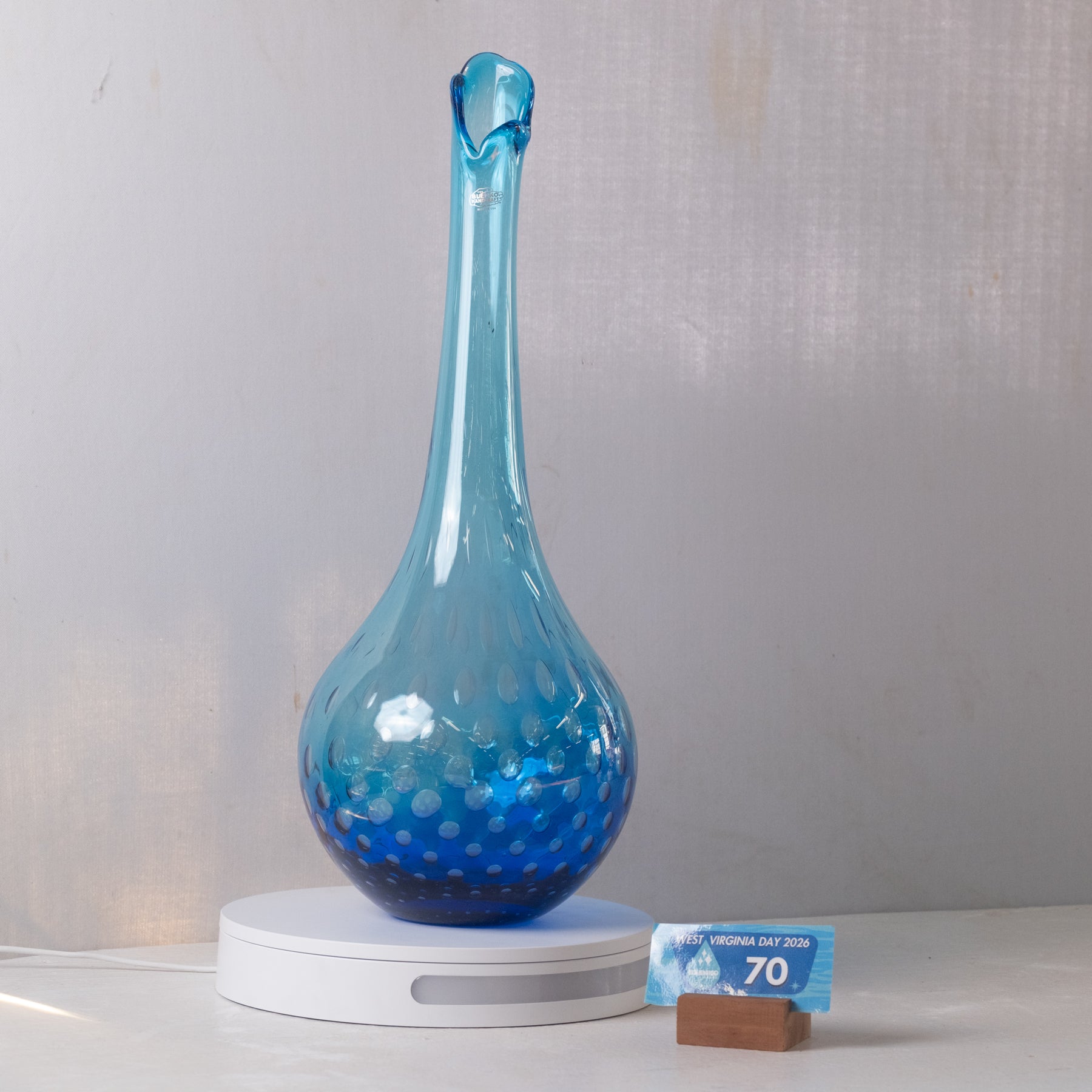 A blue glass decanter with a bulbous body and narrow neck, resting on a white circular base. The decanter has a gradient of light to deep blue with white dotted bubble patterns concentrated toward the bottom.