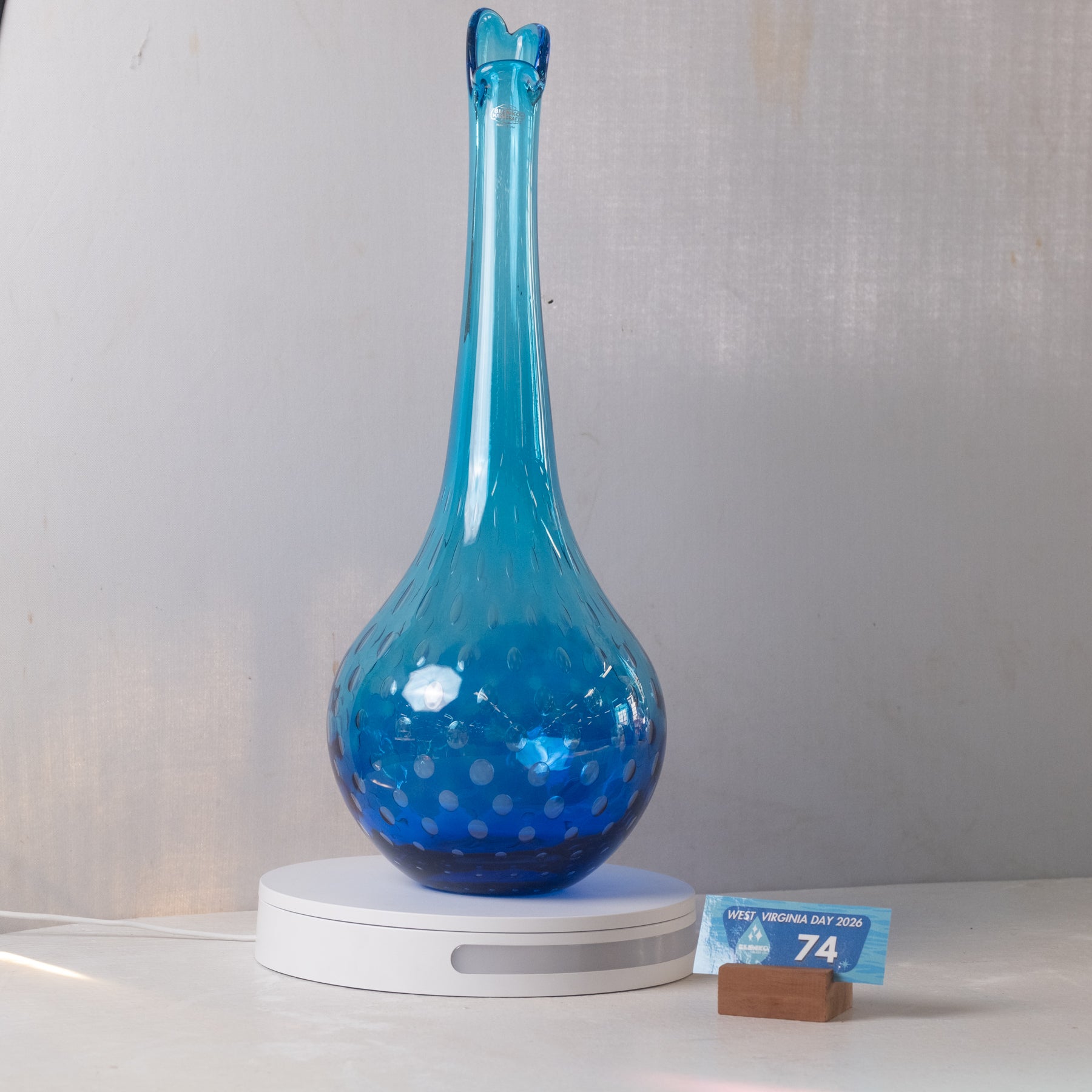 A translucent blue glass vase with a flared neck and bubble pattern, displayed on a white circular light box with a small blue price card in front, against a light gray fabric backdrop.