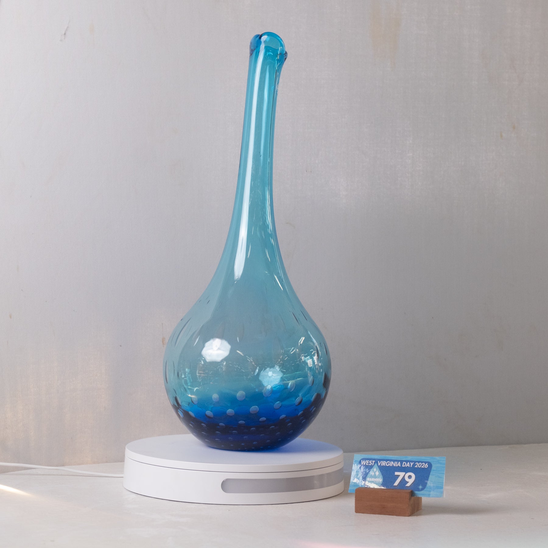 A blue glass vase with a long neck and a white circular base, displayed on a light gray background with a small price tag in front.