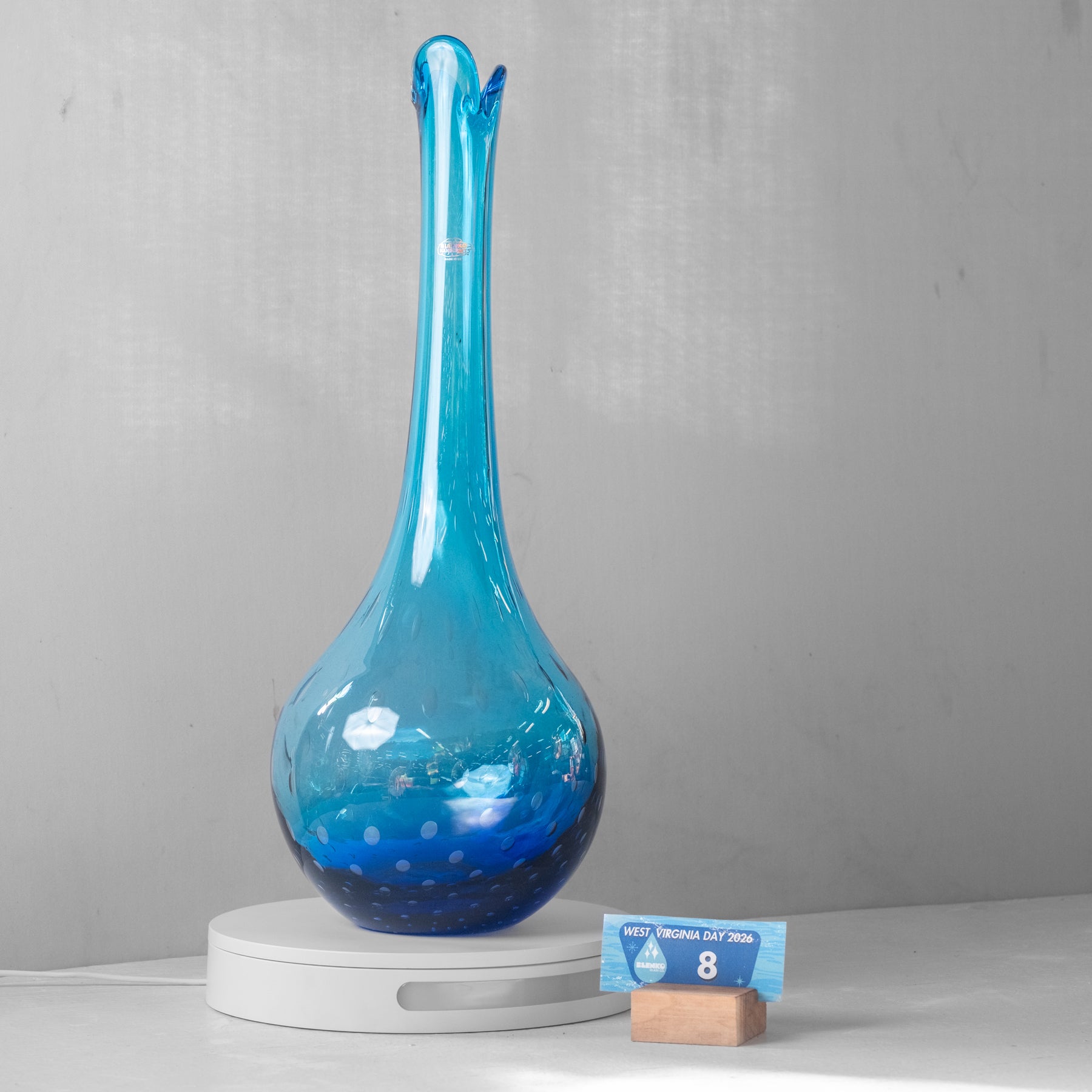A tall, slender blue glass vase with a bulbous base and a flared rim, displayed on a white circular pedestal. A small blue-and-white card on a wooden block sits beside it against a light gray wall and white surface.