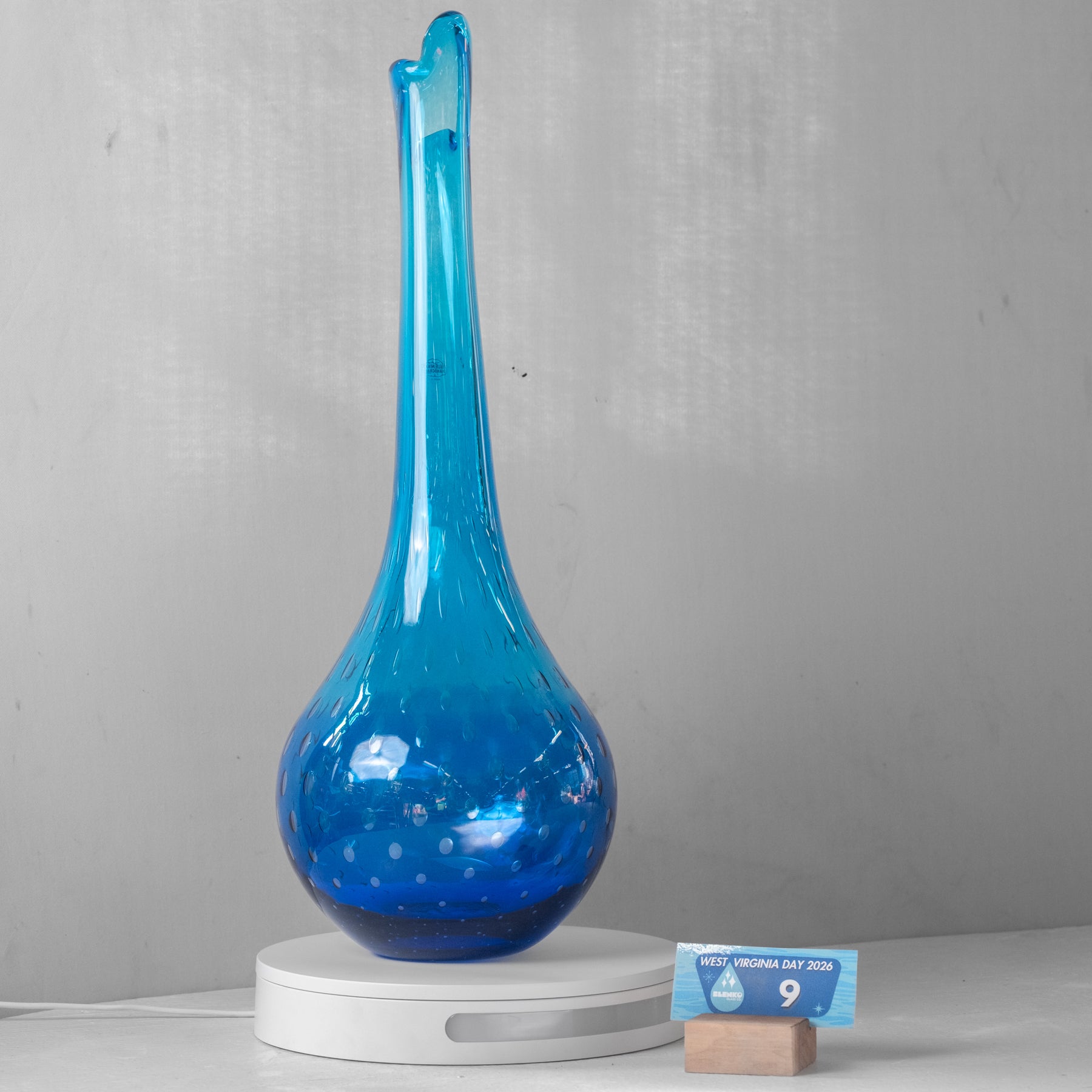 A translucent blue glass vase with a long, narrow neck and a rounded base, displayed on a white circular stand against a light gray wall. A small blue size card is placed beside the stand.