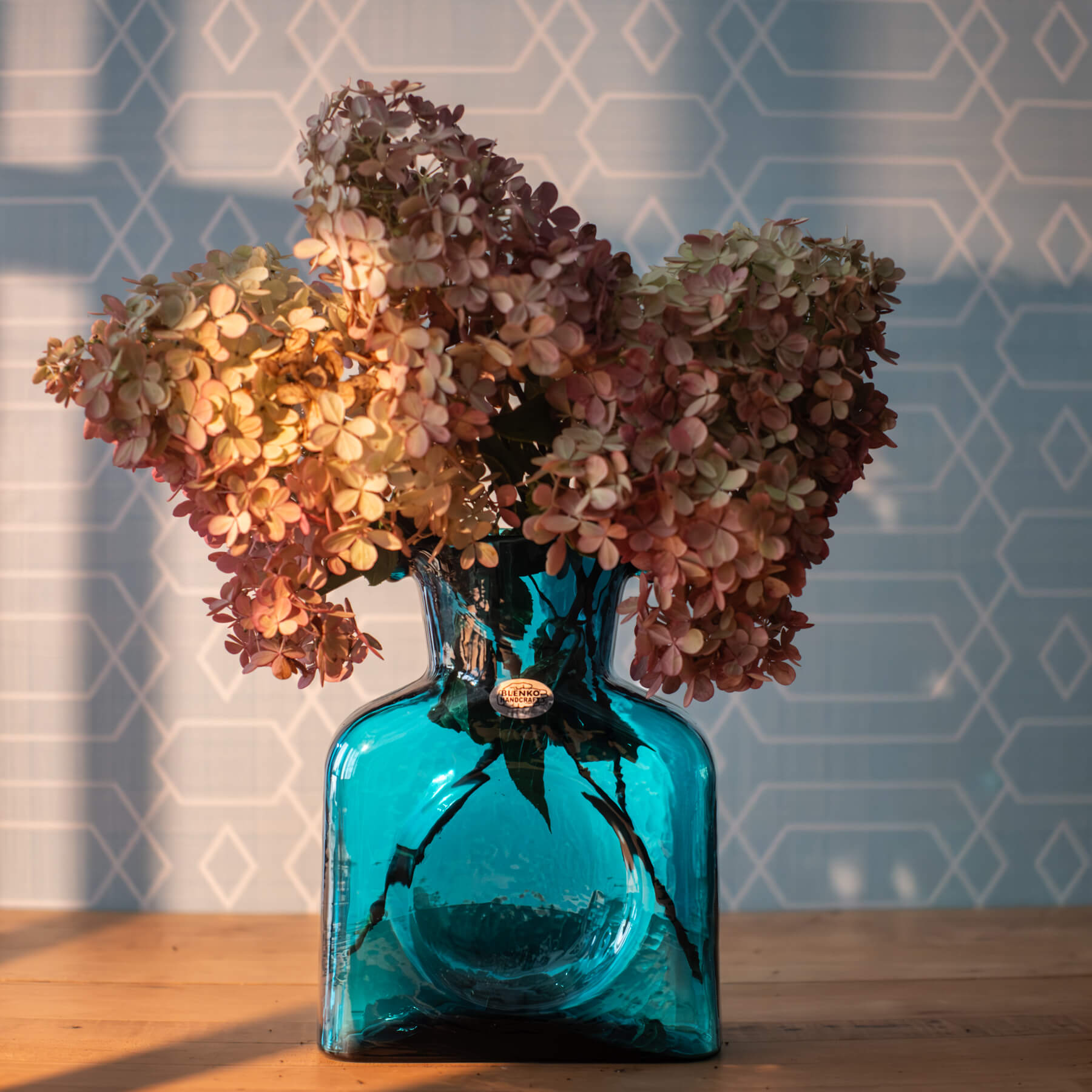 Blue glass bottle with dried flowers against a geometric-patterned wall.