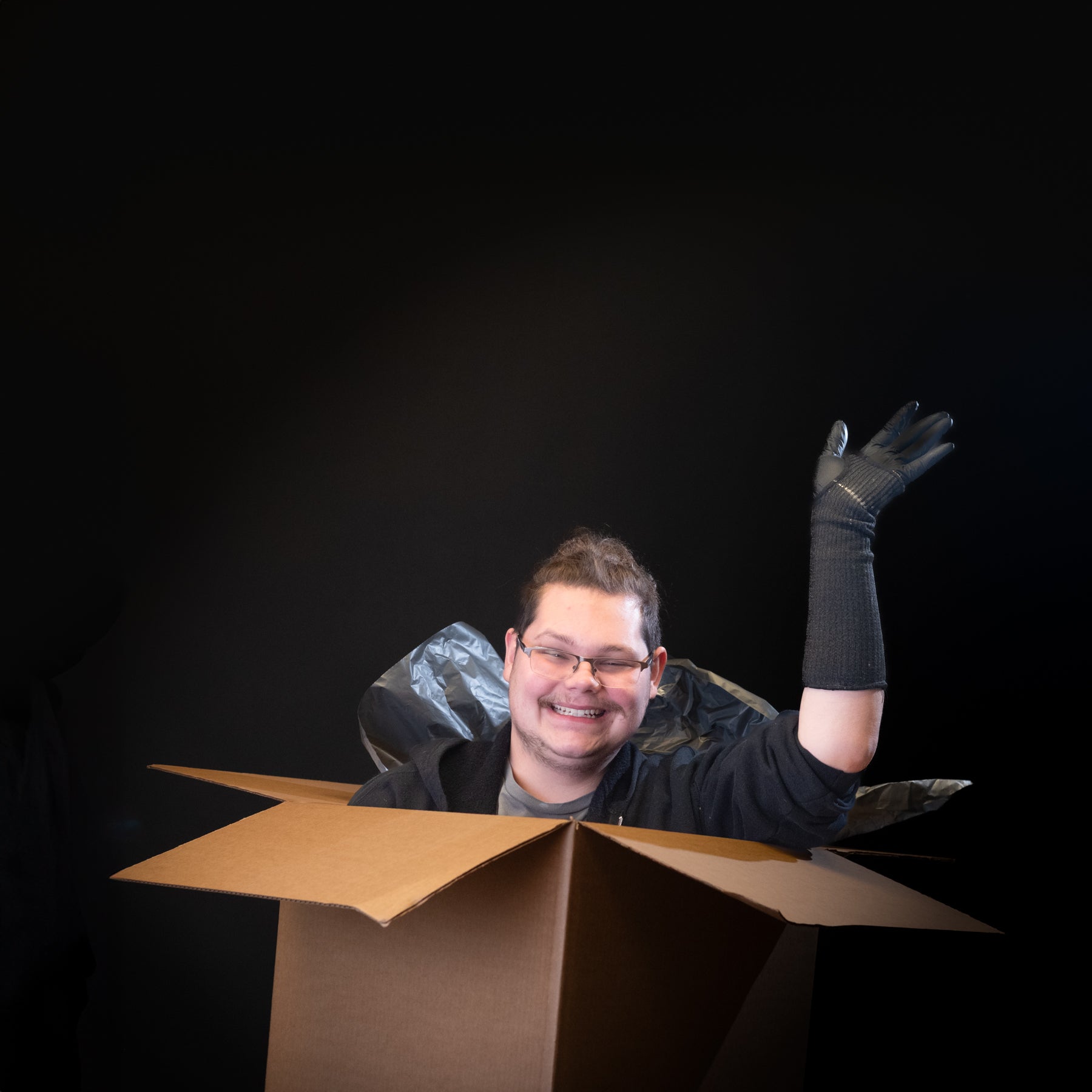 Person sitting inside a cardboard box with one arm out, wearing a black glove against a dark background