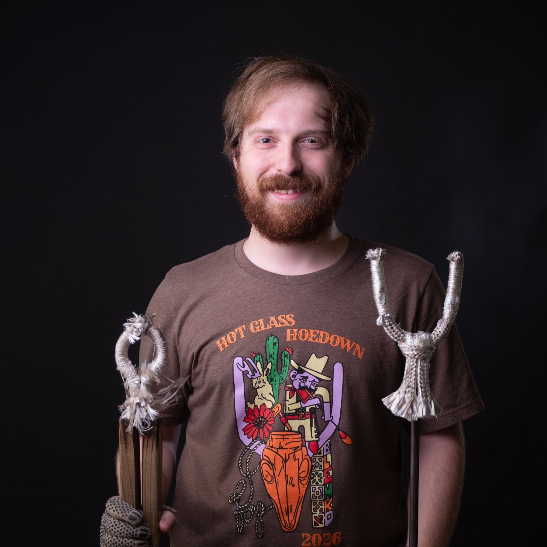 A smiling bearded man wearing a brown graphic t-shirt with 'HOT GLASS HOEDOWN 2026' printed on the front, holding a carved wooden staff topped with a skull and a white woven staff against a solid black background.