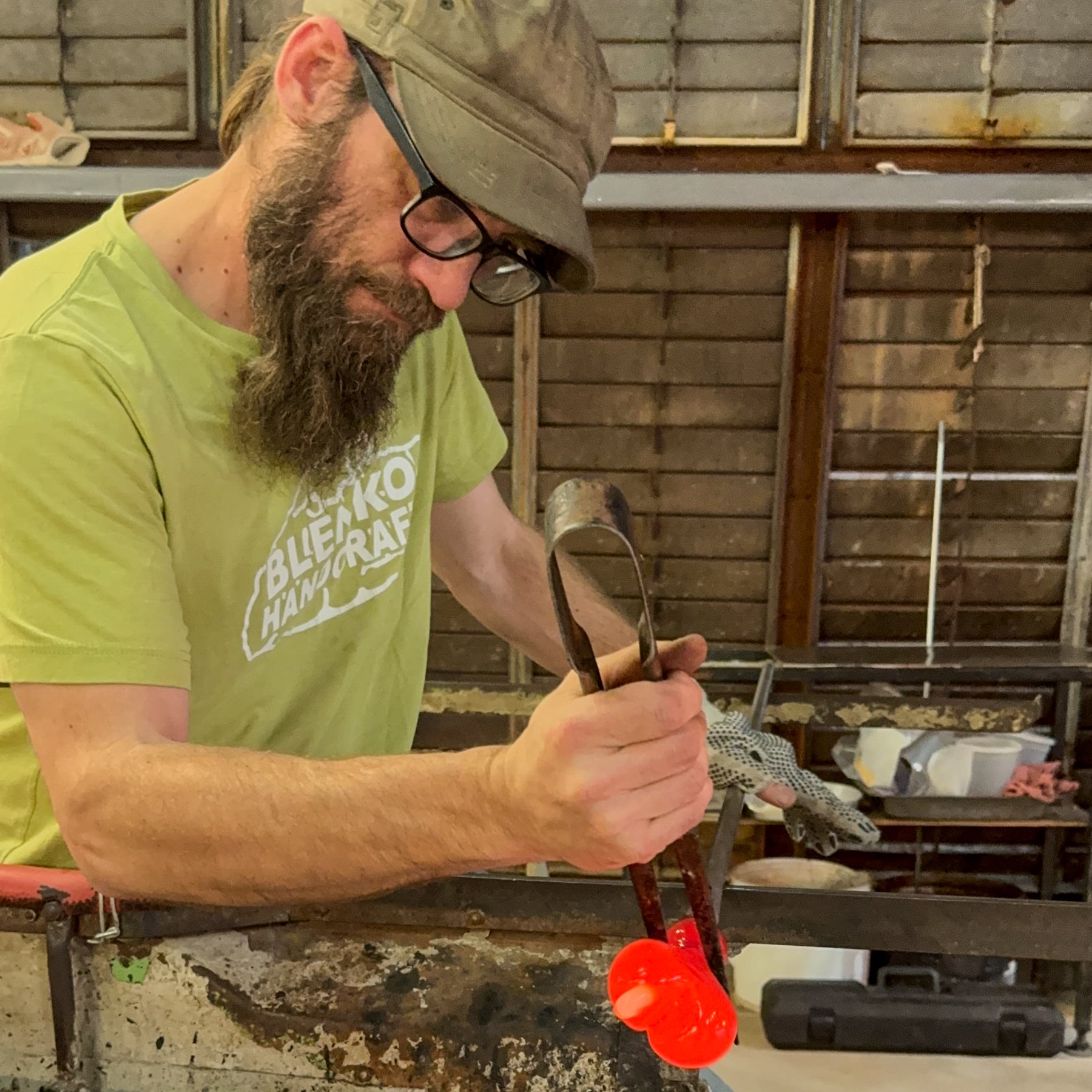 Man in a workshop holding a tool, wearing a green shirt with 'Blenko Handcraft' branding.