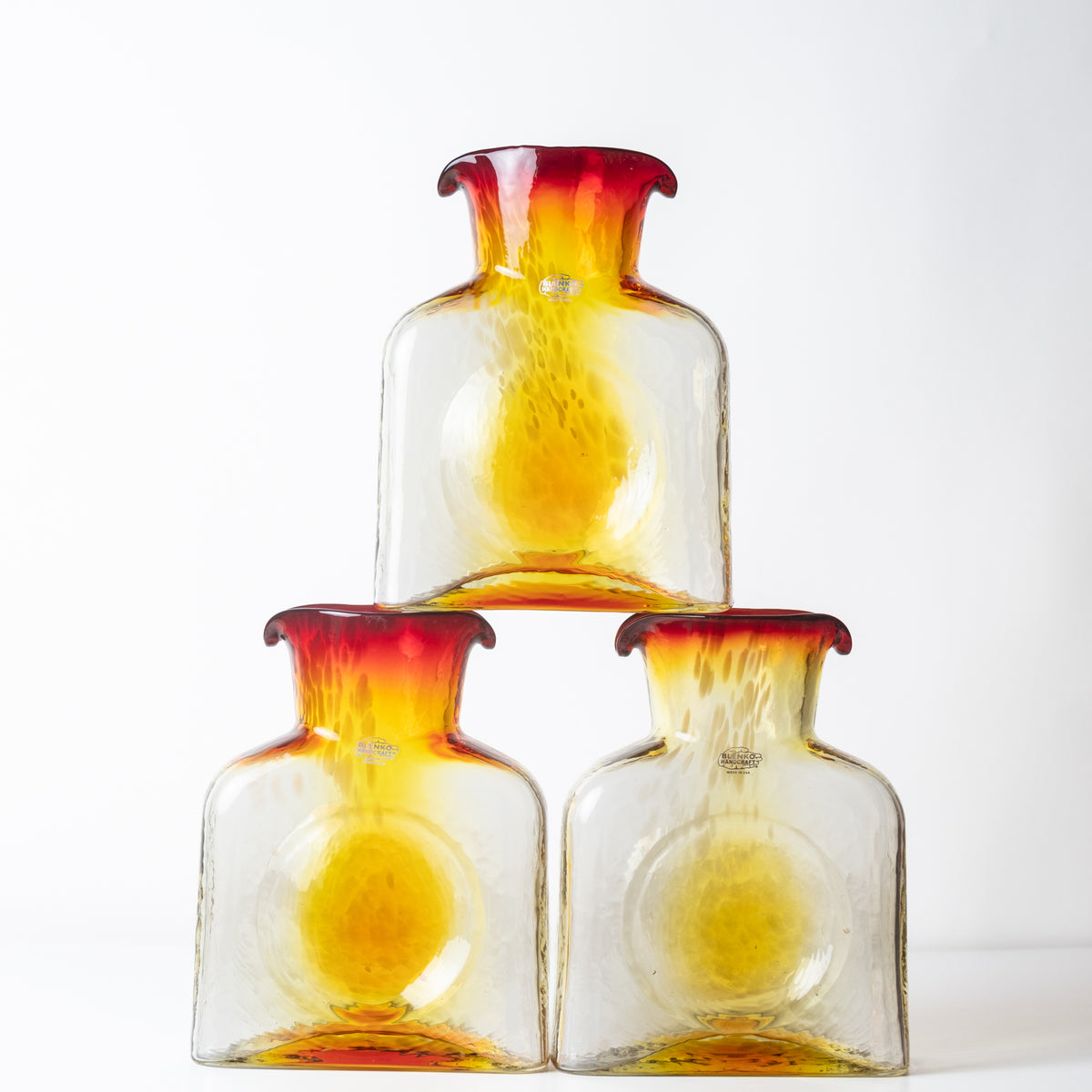 Three clear glass bottles with a yellow gradient and a dark red flared rim, stacked in a pyramid arrangement on a white background.