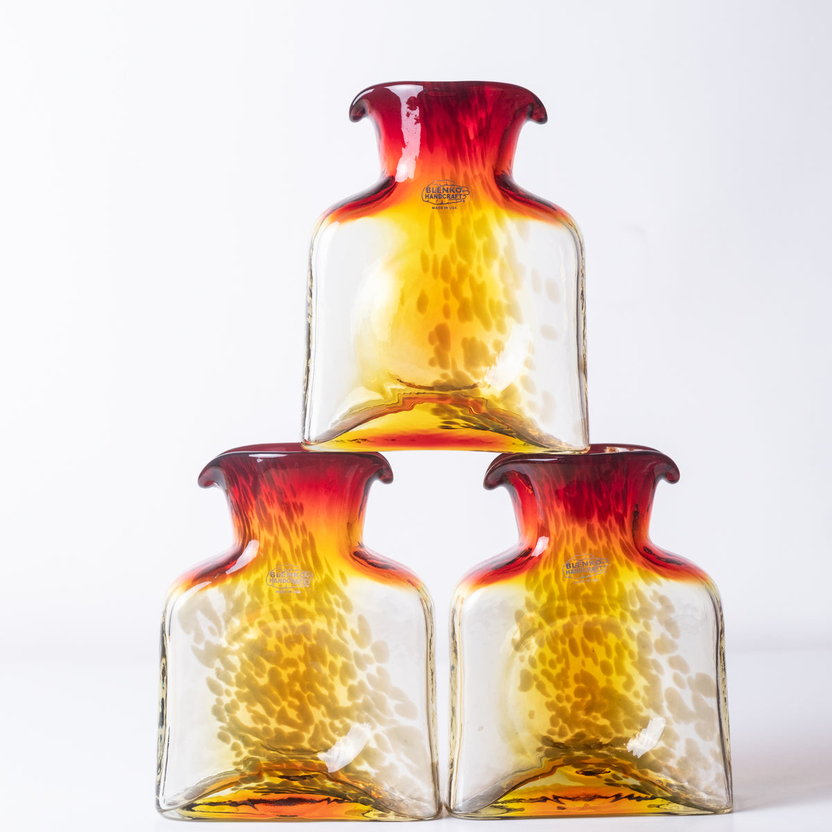 Three hand-blown glass bottles with a gradient design transitioning from deep red at the neck to translucent yellow and orange, arranged in a pyramid on a white background.