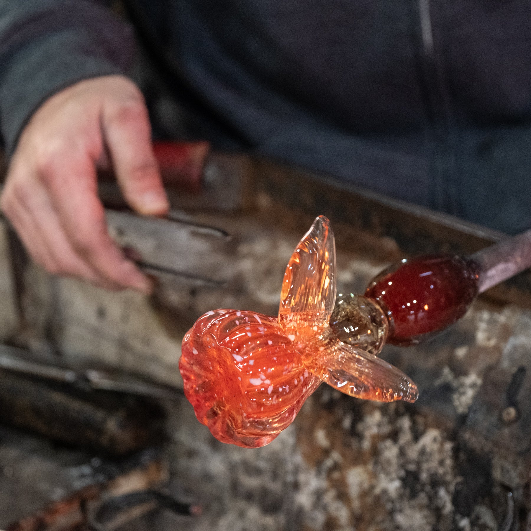 Glassblowing process with a hand holding a tool and molten glass shaped like a flower.