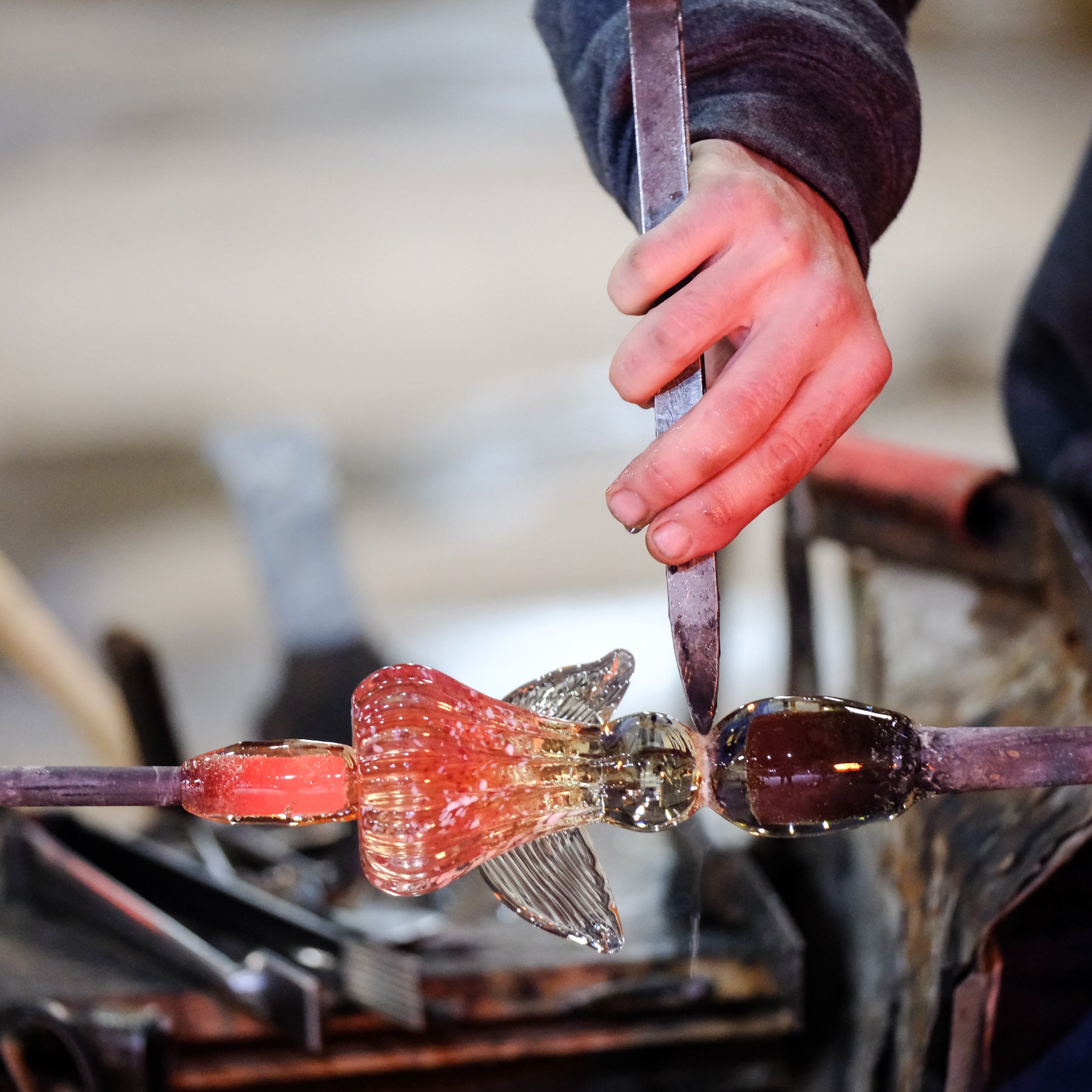 Glassblower shaping molten glass into a fish-like form with a blurred background