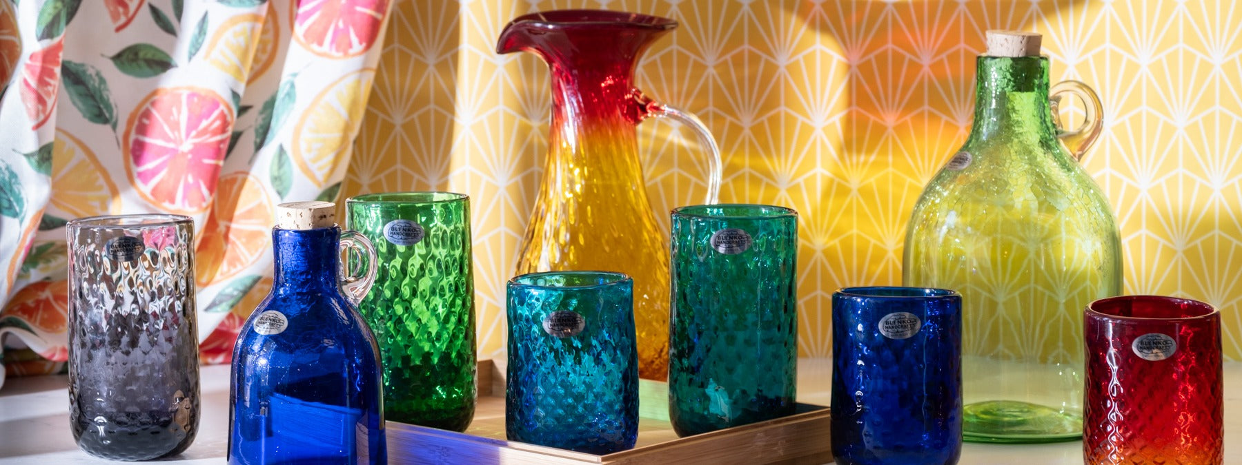 Collection of colorful glassware including bottles, tumblers, and a pitcher on a wooden surface with a patterned curtain in the background.