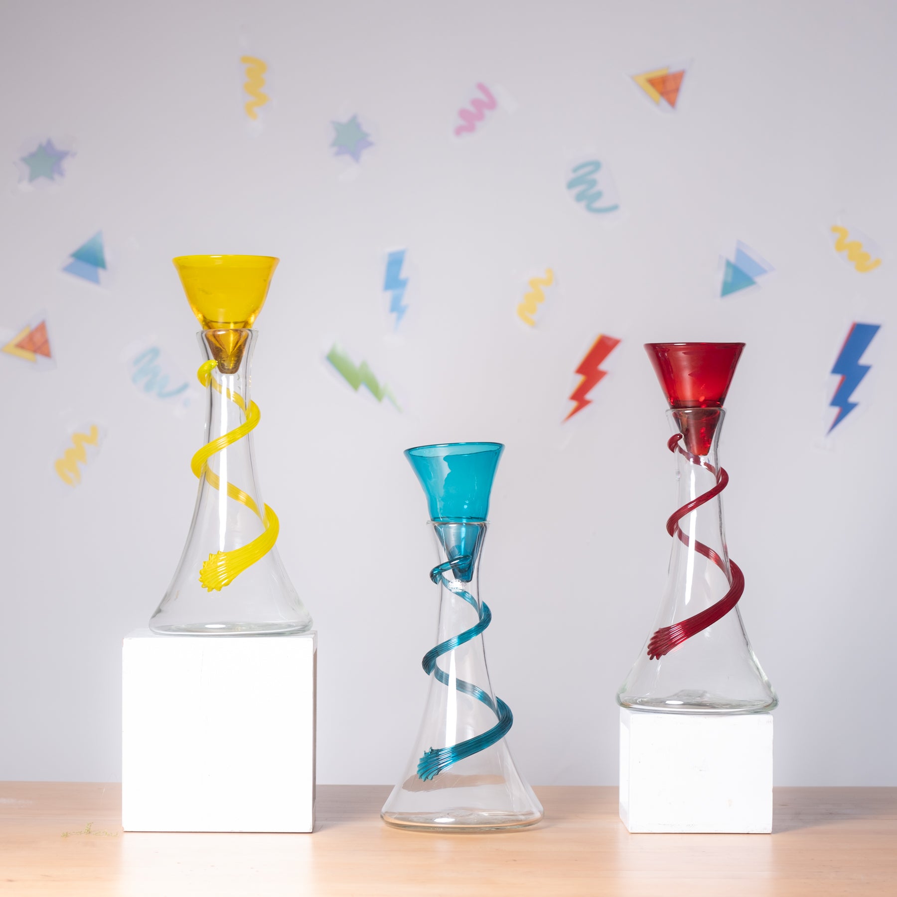 Three colorful glass decanters with spiral designs on a light gray background with abstract shapes.