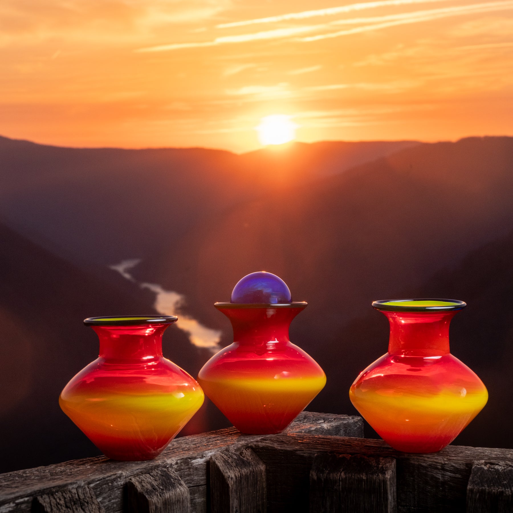 Three colorful vases on a wooden ledge with a sunset over mountains in the background