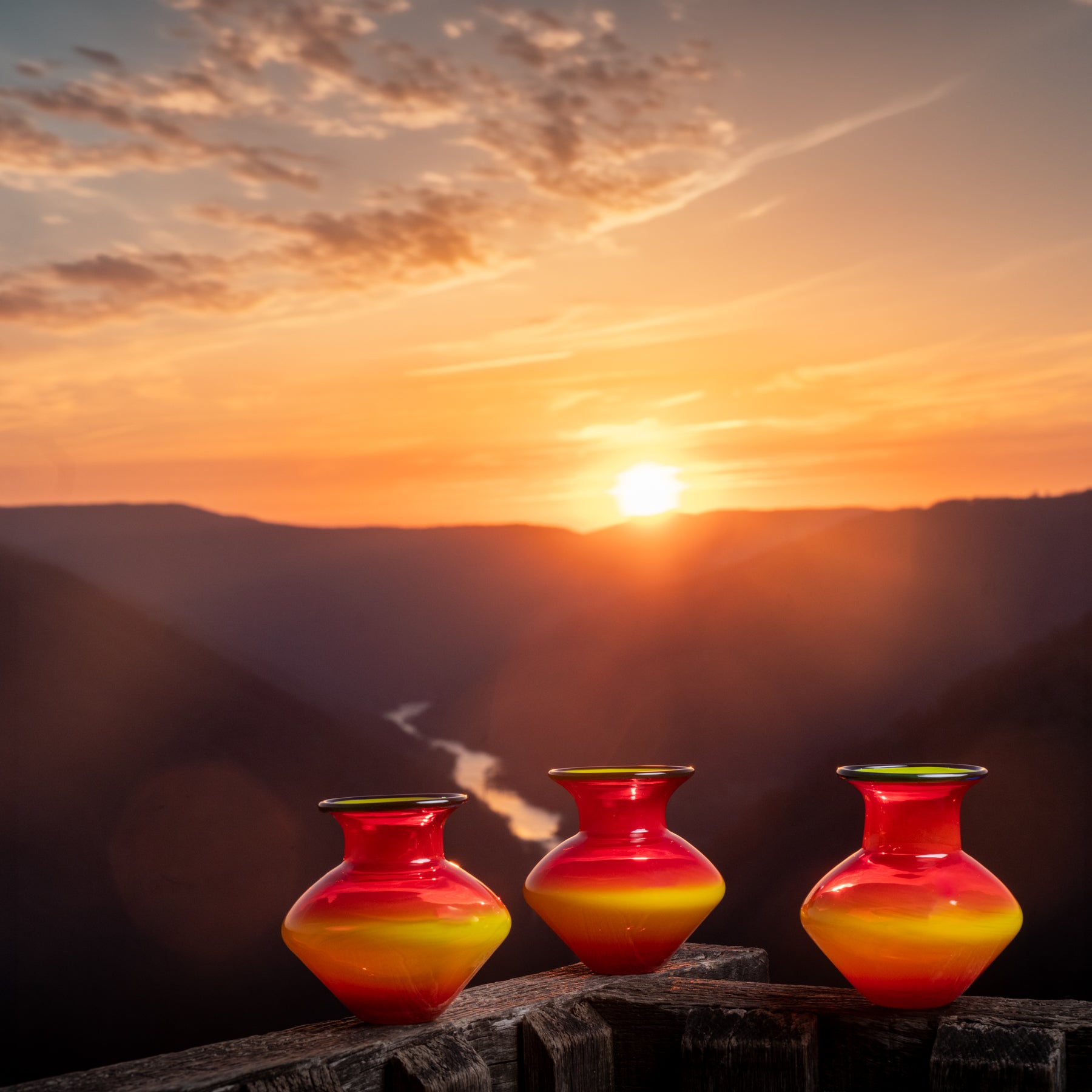 Three colorful vases on a wooden ledge with a sunset over mountains in the background