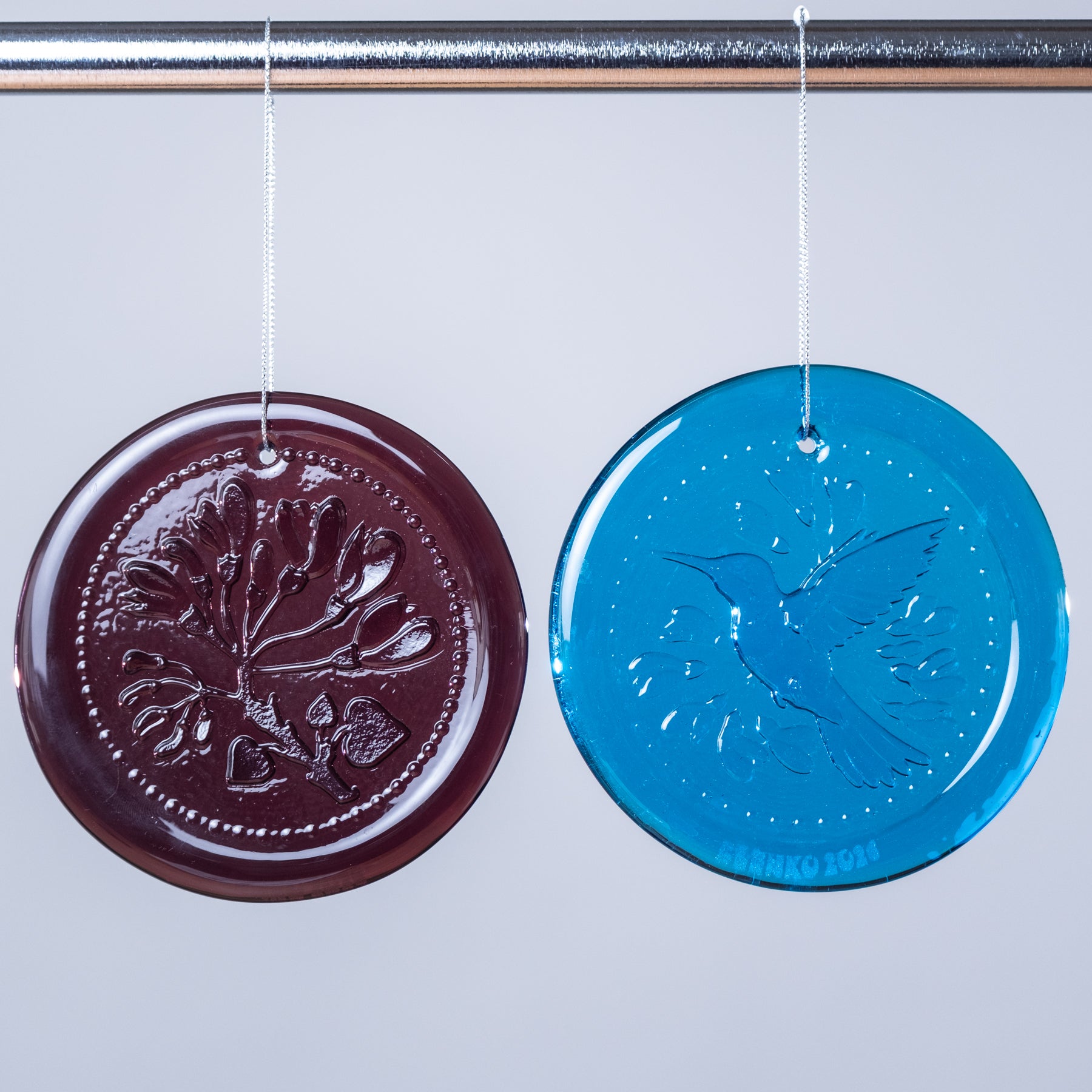 Two round glass ornaments, one brown with a tree design and one blue with a bird design, hanging against a light gray background.