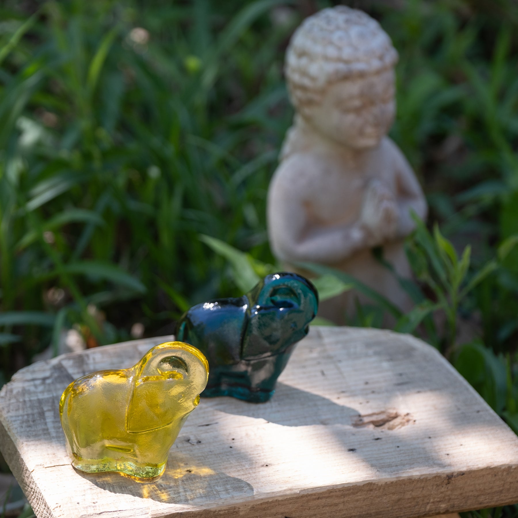 Two glass animal figurines on a wooden surface with a blurred statue of a meditating figure in the background.