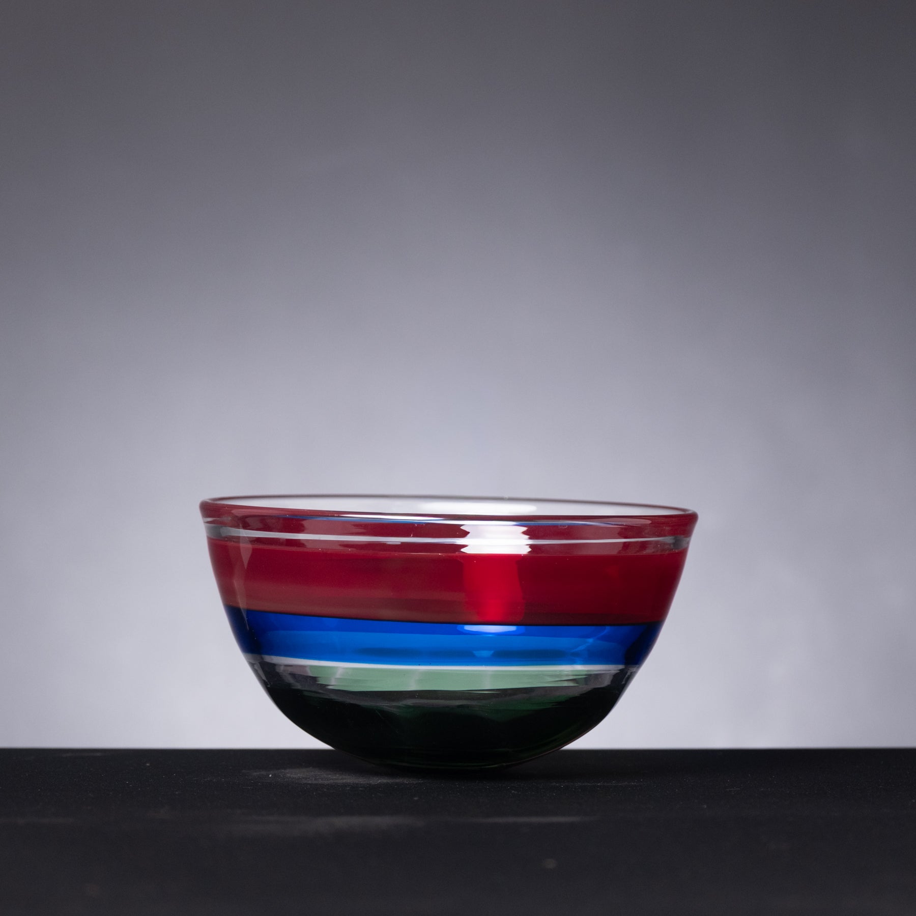 Colorful glass bowl with red, blue, and green stripes on a dark surface and gray background