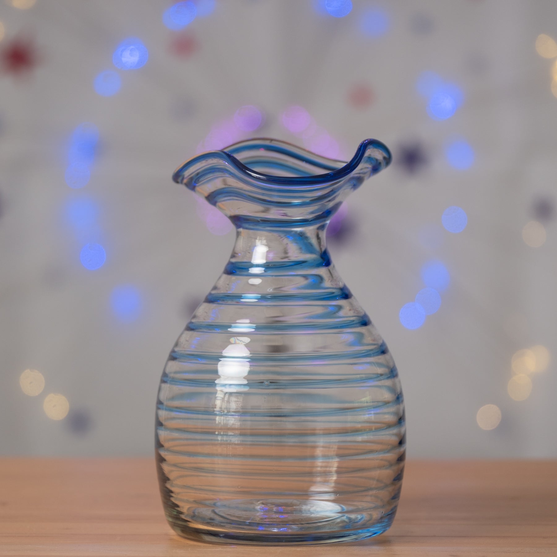 Clear glass vase with blue ribbed design on a wooden surface with blurred colorful lights in the background