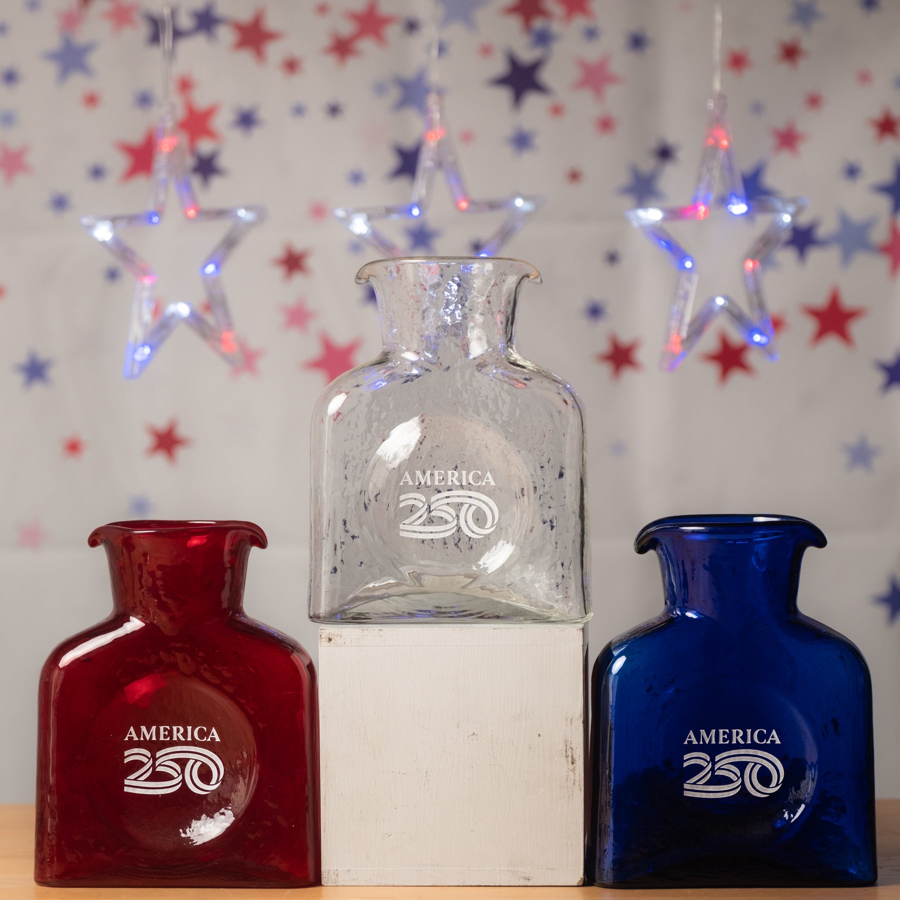 Three glass bottles, red, white, and blue, with 'America 250' engraving on a star-patterned background.