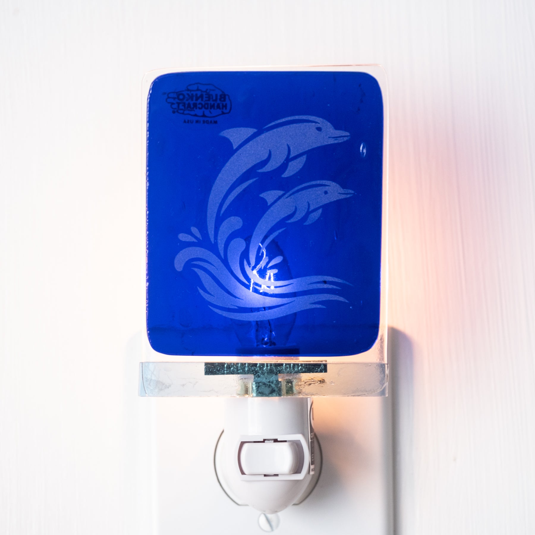 A blue dolphin-themed night light plug-in with a white switch, shown plugged into a wall outlet against a white wall.