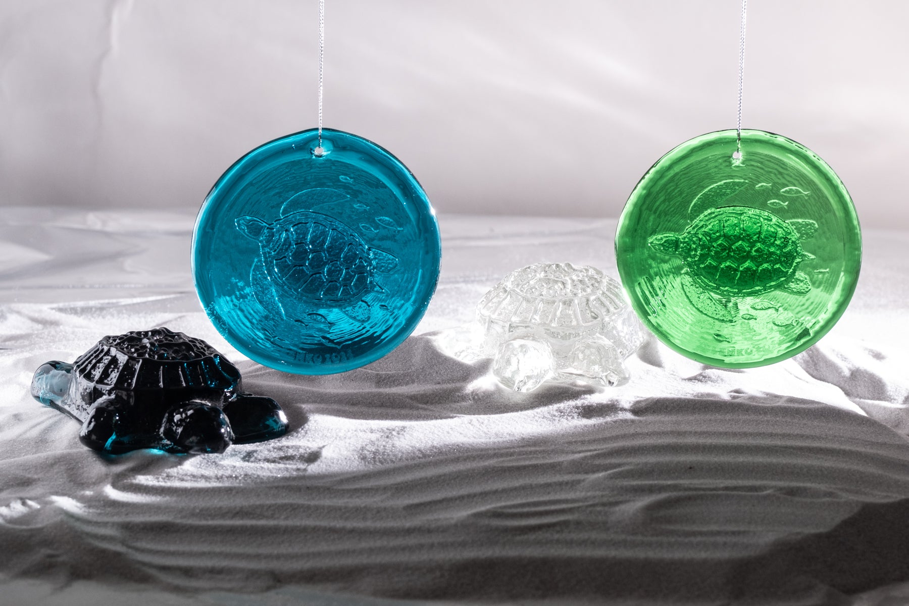 Three hanging turtle-shaped glass ornaments in blue, green, and black, displayed on white fabric with a white draped backdrop.