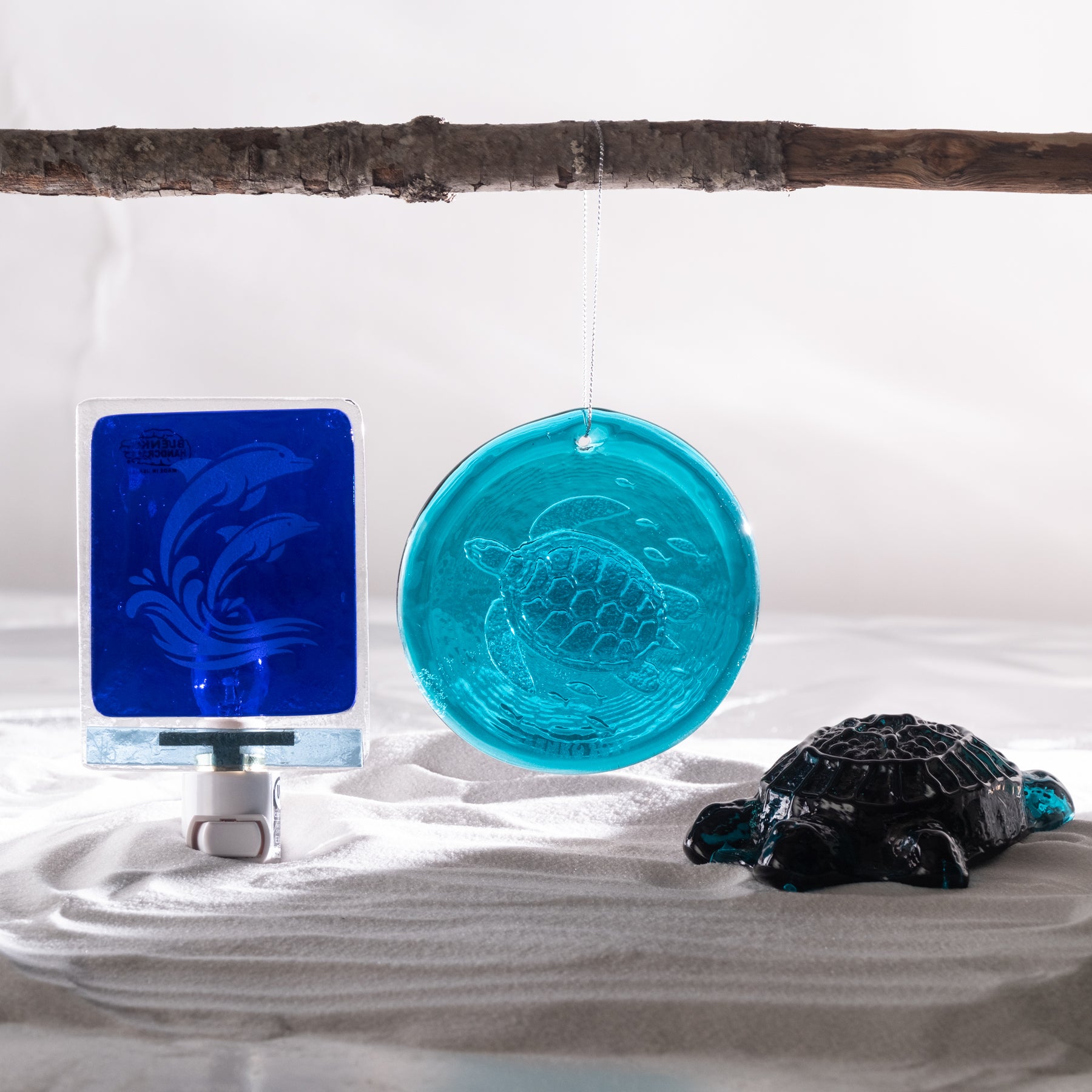 A decorative glass ornament shaped like a sea turtle hangs from a branch above white sand, next to a blue square glass piece on a small stand and a black turtle figurine.