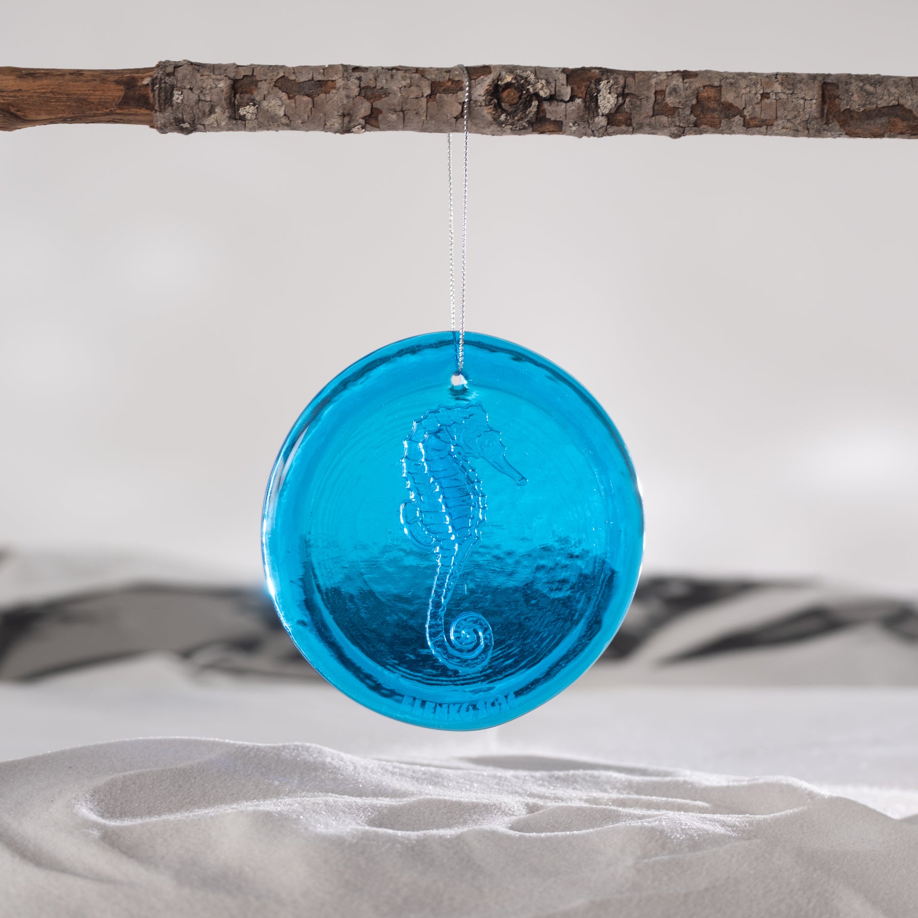 A round, translucent blue glass ornament featuring an embossed seahorse design, hanging from a thin string suspended from a rustic branch over a white sandy surface.