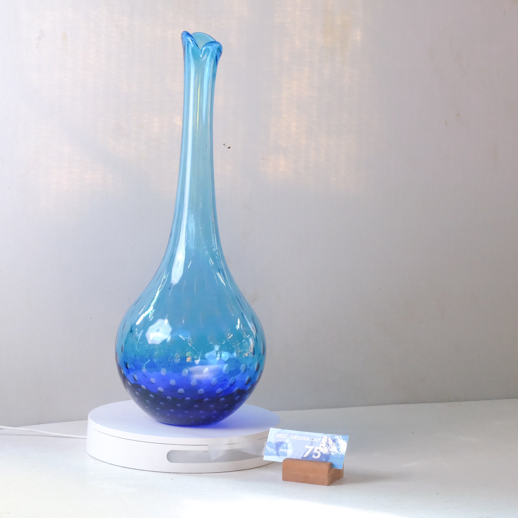 A blue glass vase with a long neck and rounded base, sitting on a white circular charging or display base against a light gray wall. A small price tag card is placed in front of the base.