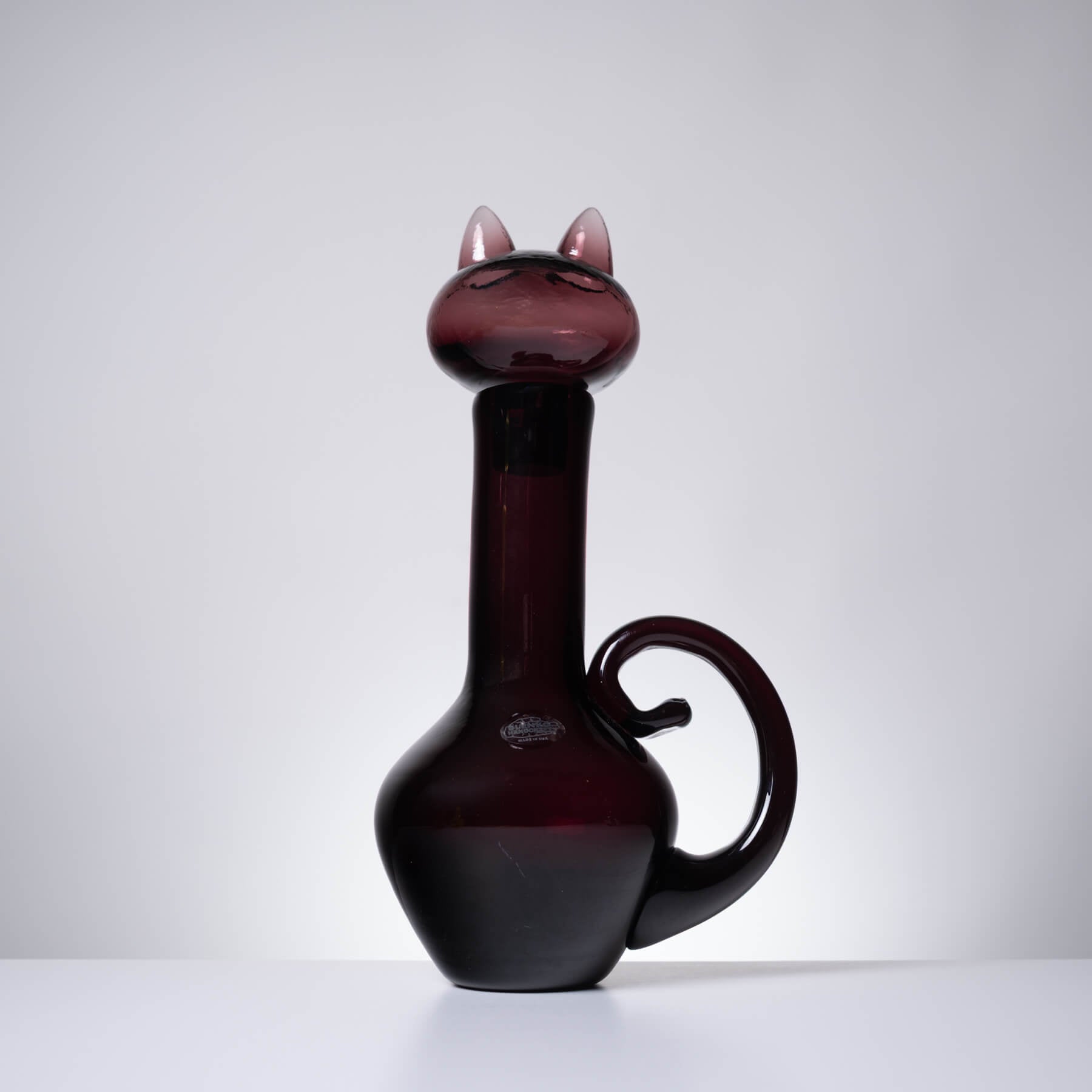 A dark burgundy glass decanter shaped like a cat, featuring pointed ears on the neck, a curved handle, and a glossy finish, set against a neutral gray background.