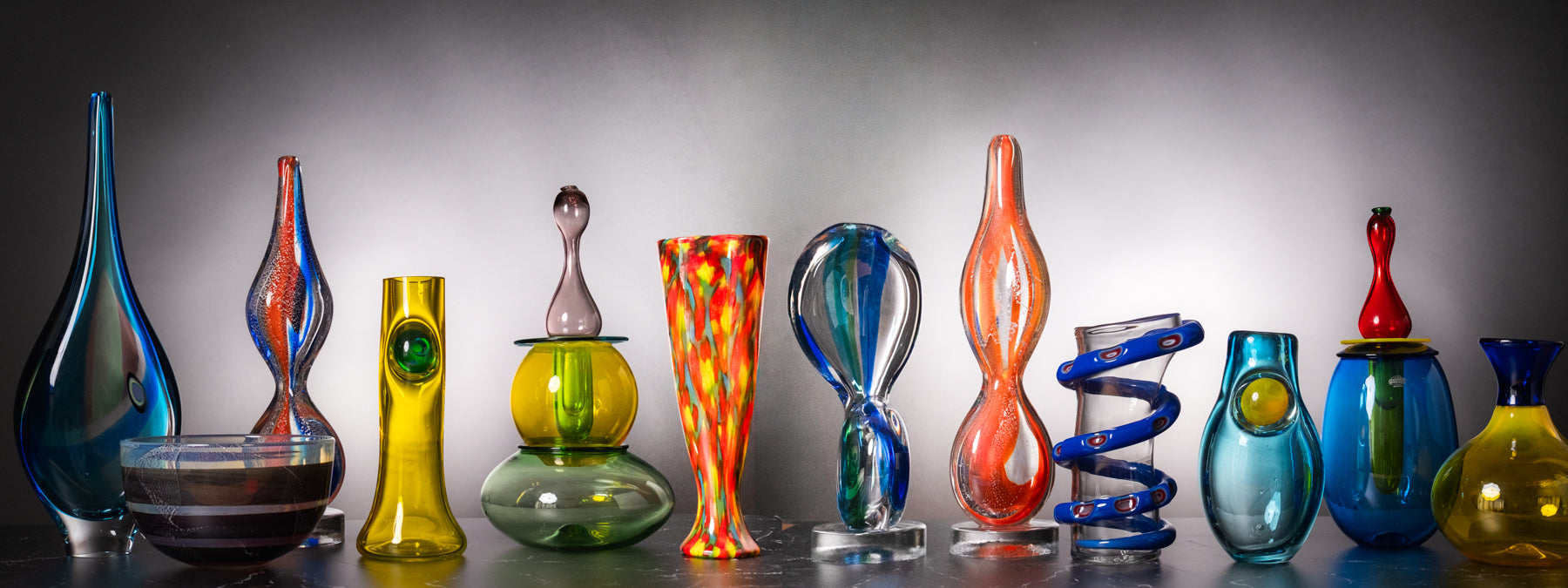 A collection of colorful hand-blown glass vases and sculptures displayed on a reflective surface against a gray gradient studio background.