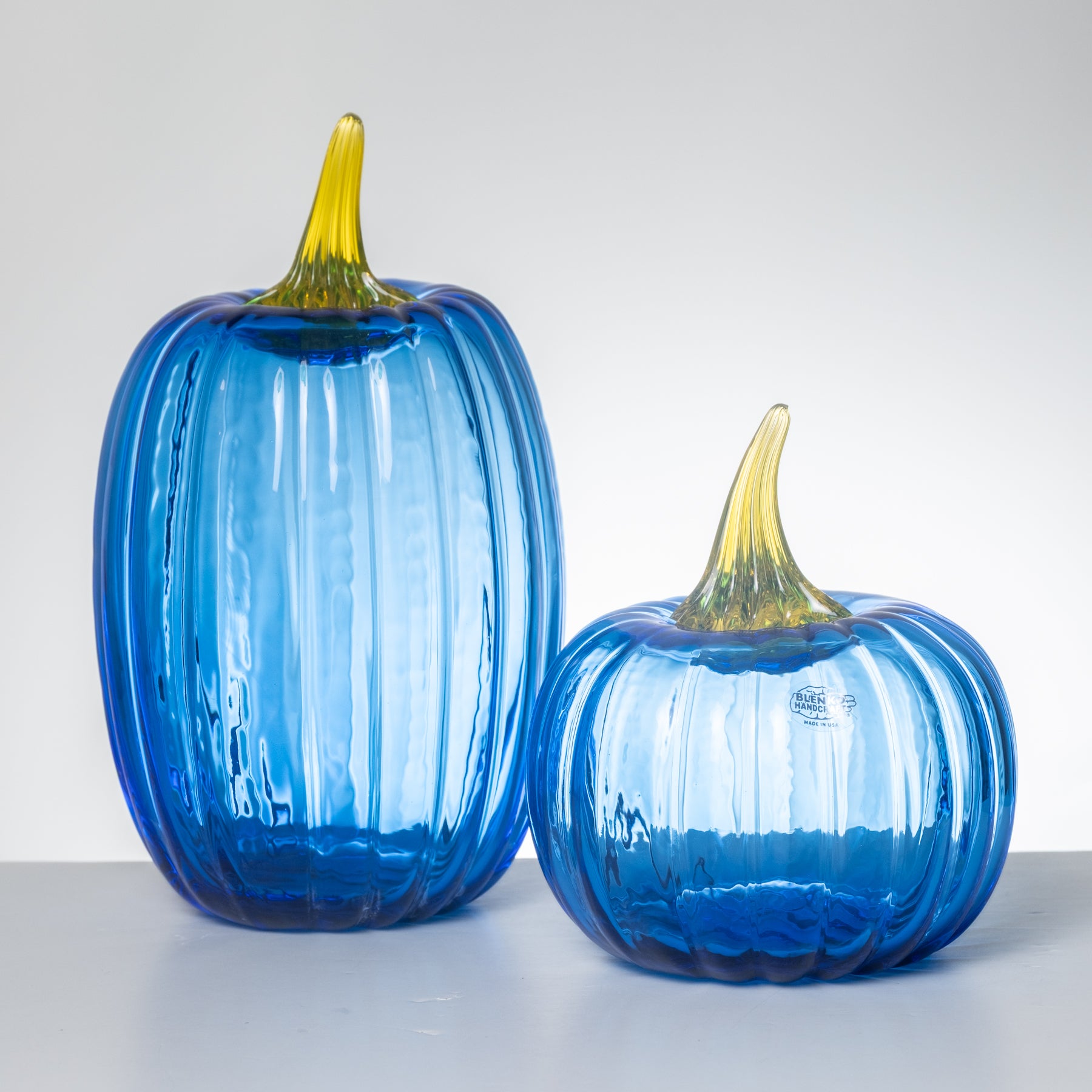 Two ribbed glass pumpkins in translucent cobalt blue with yellow stems, displayed on a white surface against a light gray background.