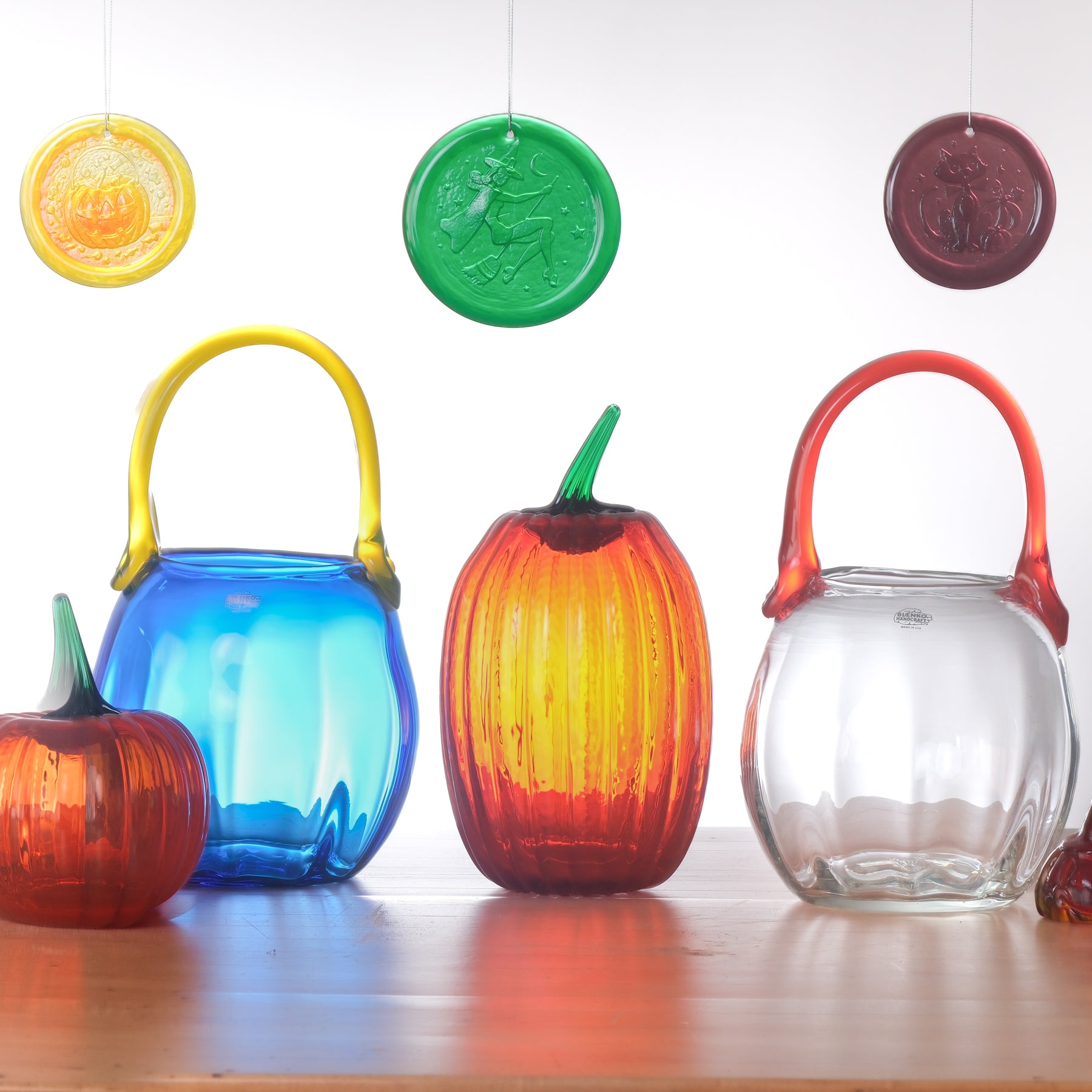 Four decorative glass pumpkins and lanterns in assorted colors are arranged on a wooden surface against a white background, with three matching hanging circular glass ornaments above. The set includes translucent orange, blue, red, and clear glass designs with ribbed pumpkin bodies and arched handles.