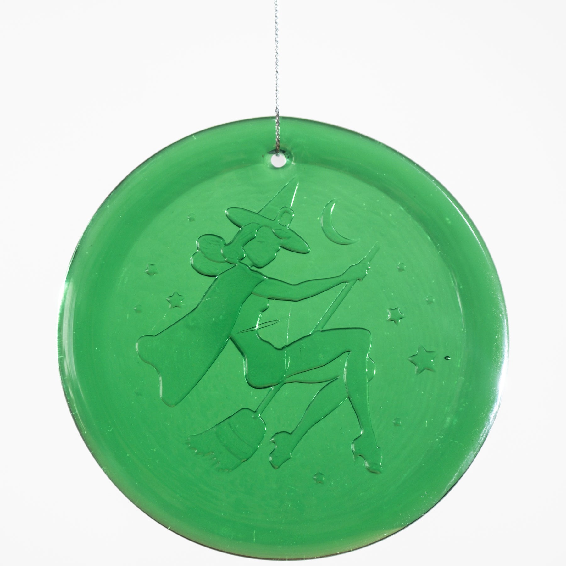 A round green glass hanging ornament featuring an embossed design of a witch riding a broomstick under a crescent moon, suspended by a thin string against a white background.