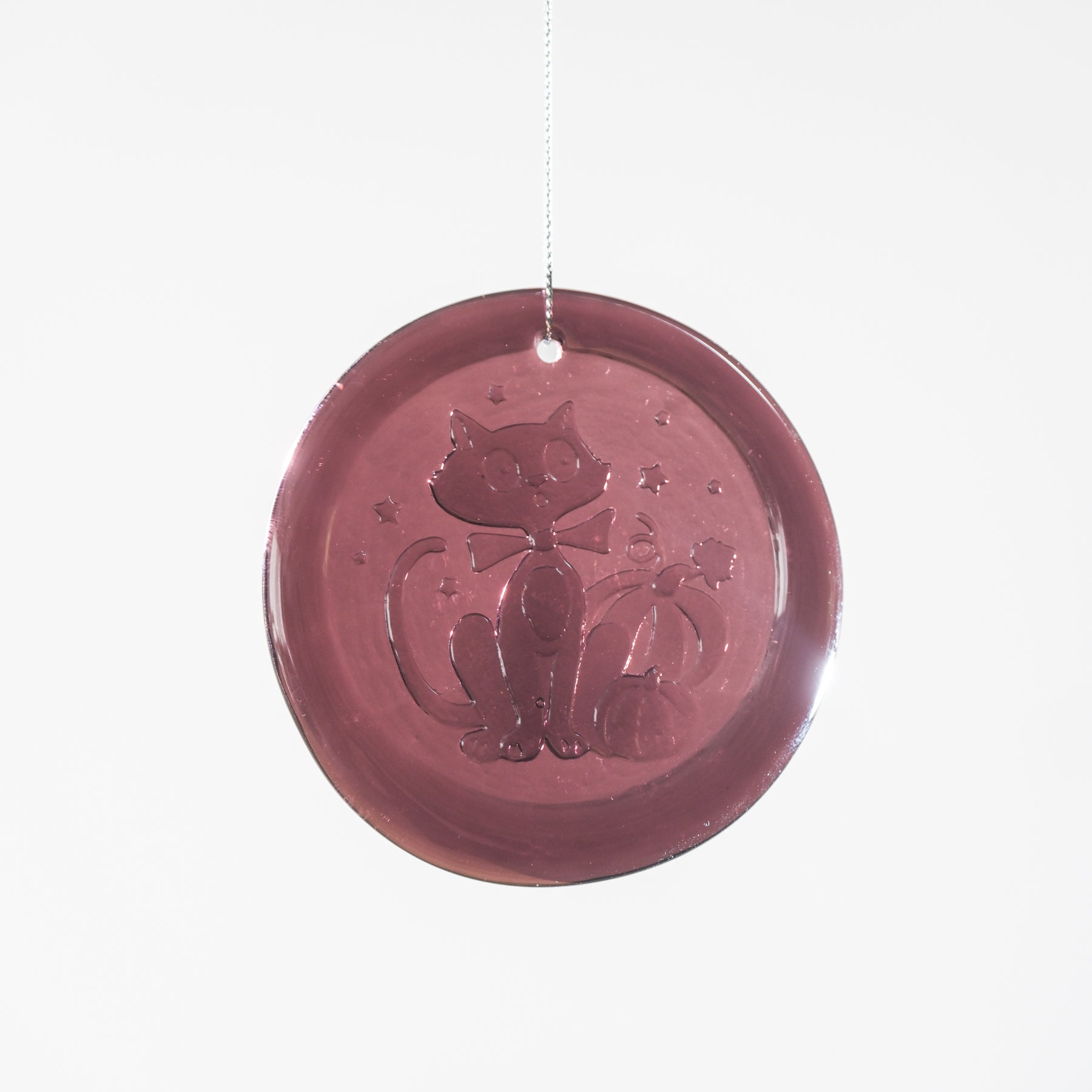 A round, translucent dusty rose glass ornament featuring an embossed design of a cat wearing a bow tie and holding a small pumpkin, suspended by a thin glass string against a plain white background.