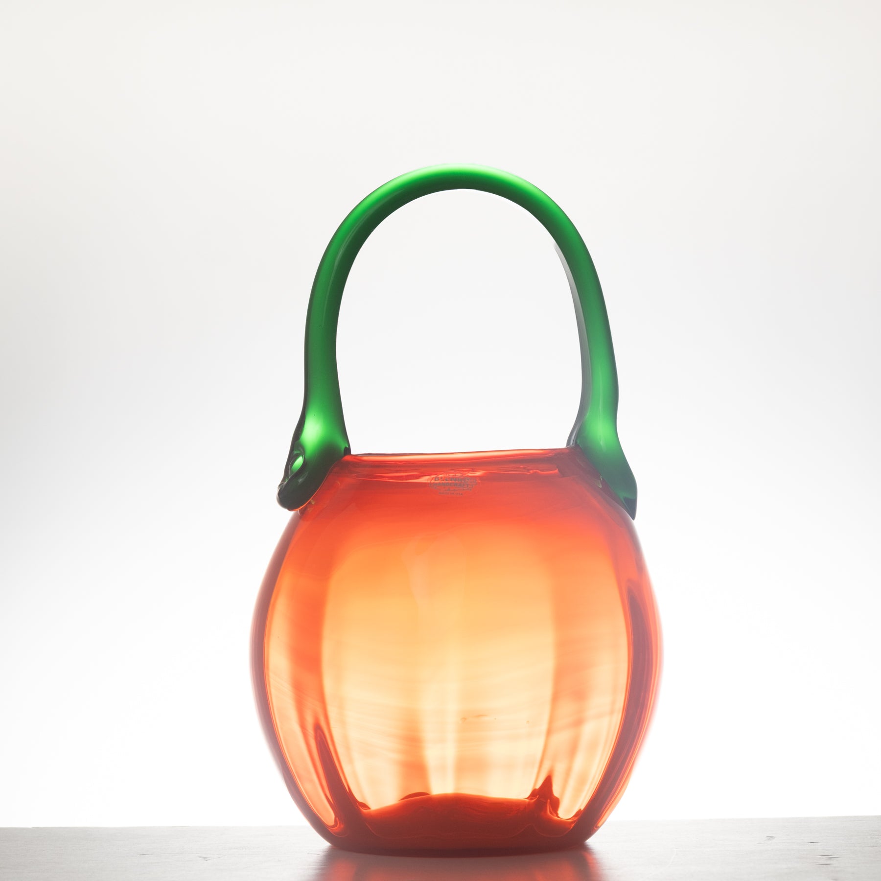 A orange glass handbag with a rounded, fish-shaped body and a green handle, shown on a neutral light gray background.