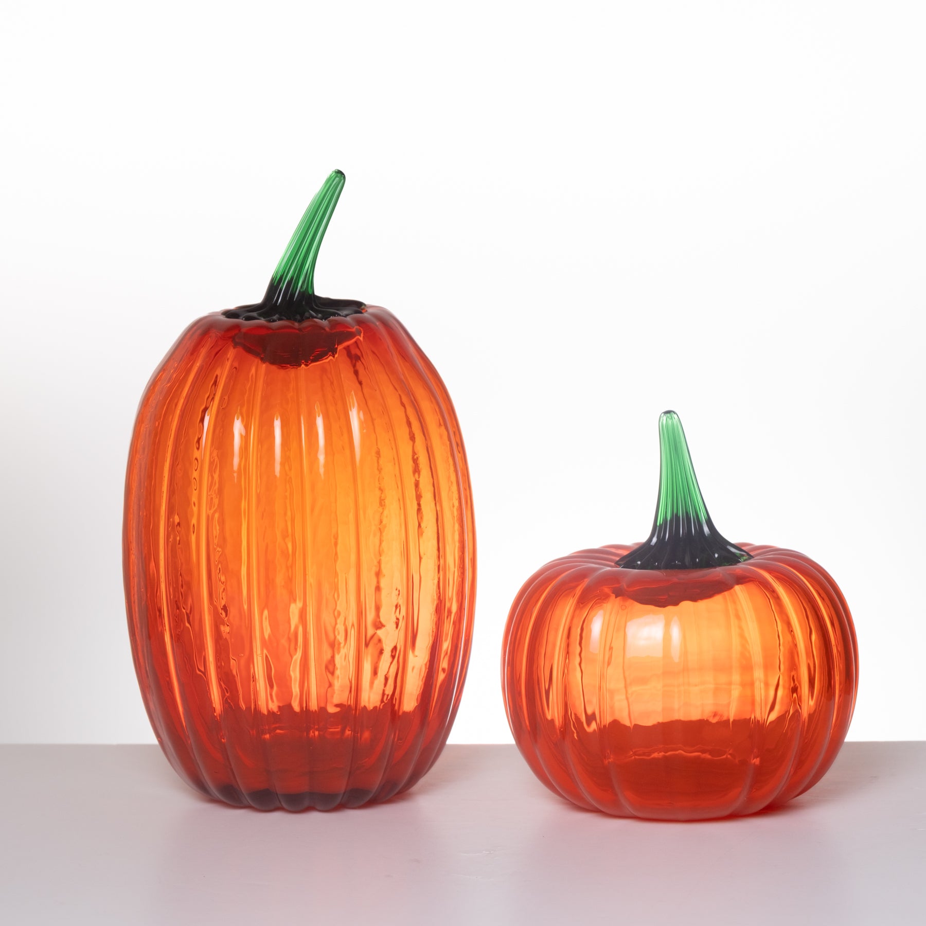 Two ribbed glass pumpkin-shaped vases in orange-red tones with green stems, displayed on a white surface against a white background.