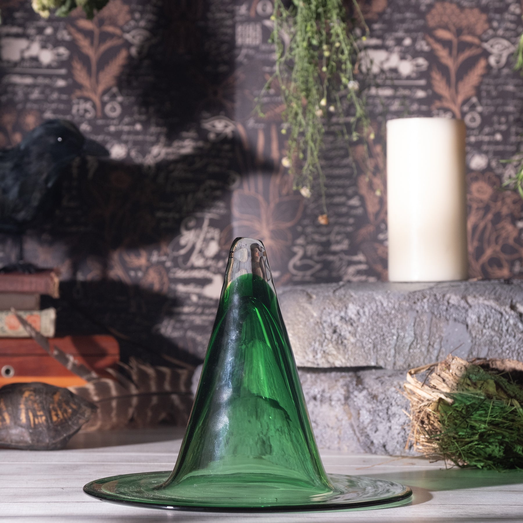 A green glass witch hat with a wide brim, displayed on a light-colored surface against a dark, moody background with a raven, books, a candle, and dried foliage.