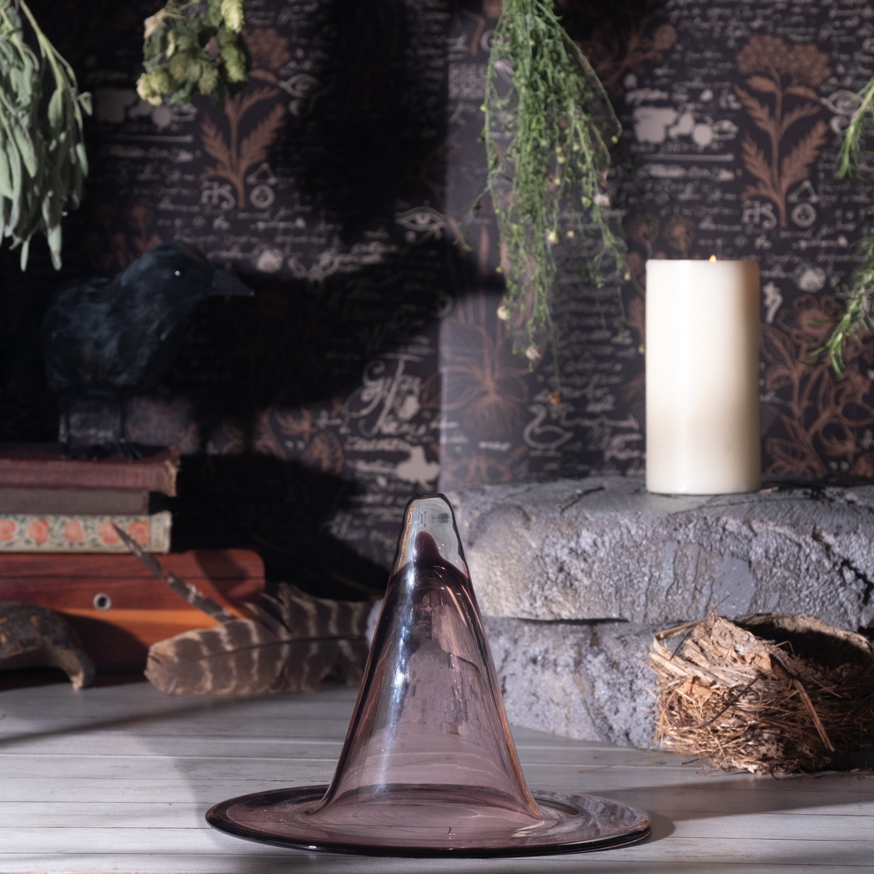 A translucent smoky purple glass conical object, resembling a witch hat or decorative goblet, displayed on a stone surface in a moody styled setting with a candle, books, dried foliage, and a dark patterned backdrop.