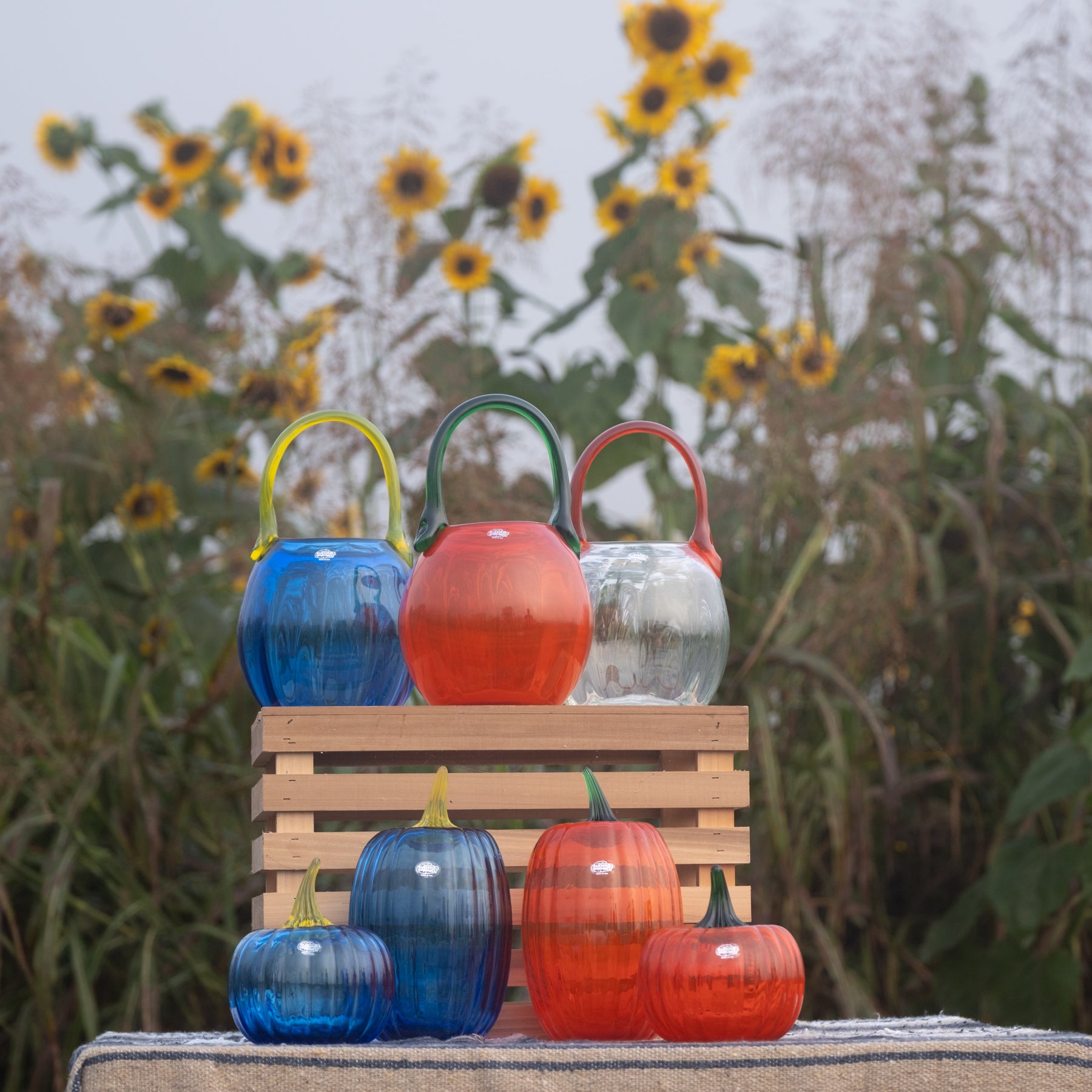 A set of colorful ribbed glass decorative pumpkins and gourds displayed on a wooden crate outdoors, with sunflowers blurred in the background.