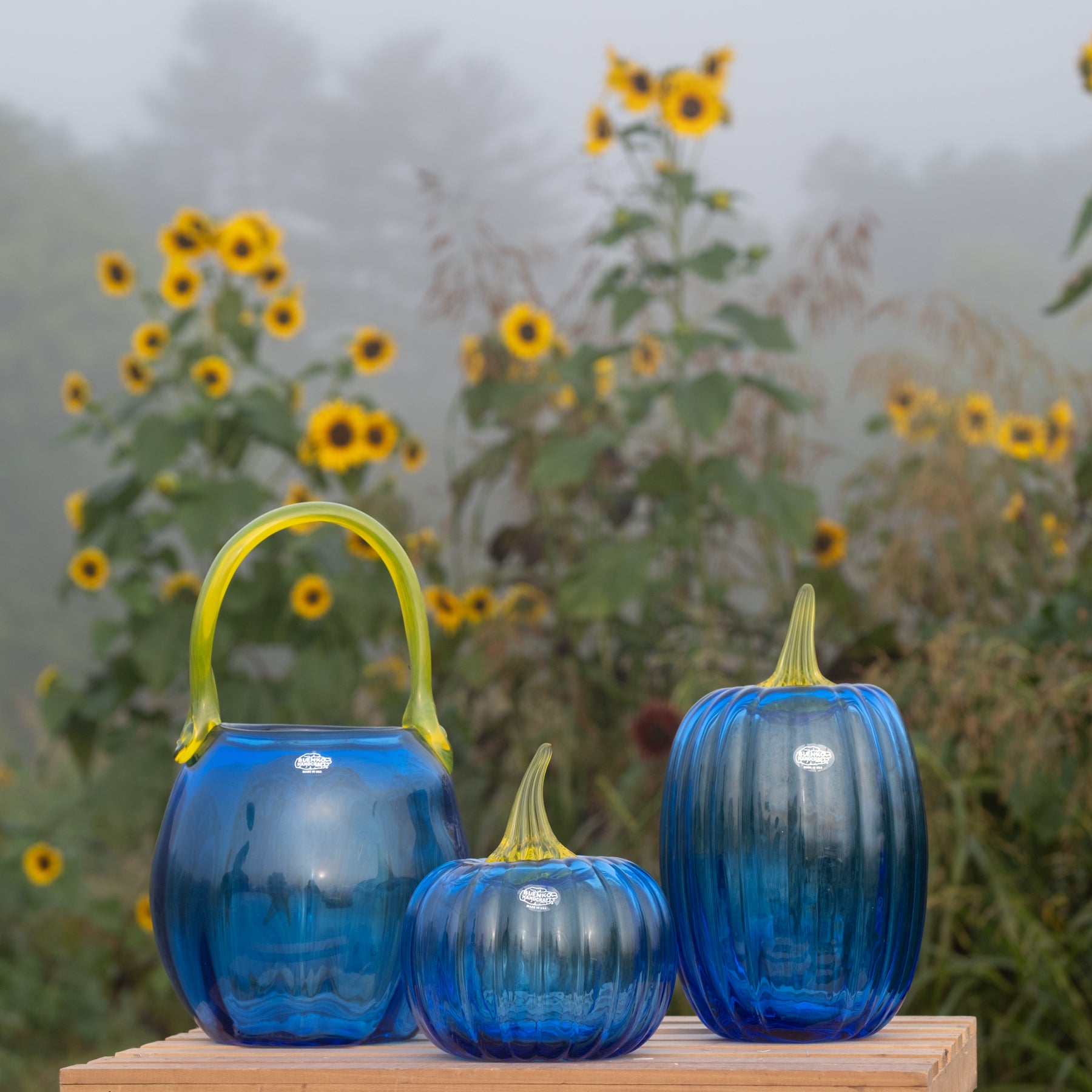 Three ribbed cobblestone-style glass vases in shades of blue with green stems and handles, displayed on a wooden surface with a blurred background of sunflowers and mist.
