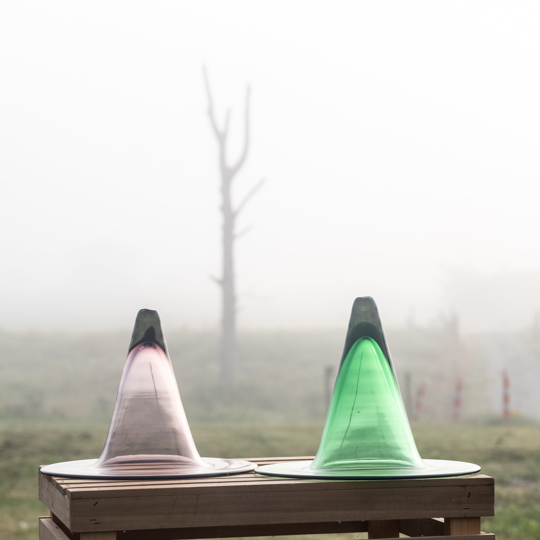 Two translucent cone-shaped glass vessels, one pink-tinted and one green-tinted, displayed side-by-side on a wooden table in a misty outdoor setting.