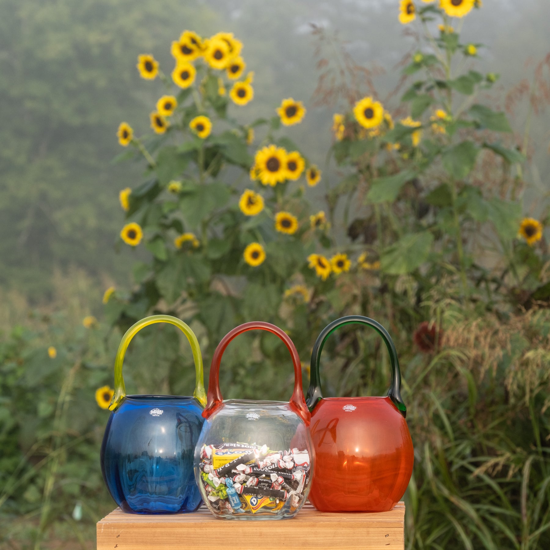 Three colorful glass teapot-shaped containers with handles, standing on a wooden block outdoors with a blurred background of sunflowers and greenery.