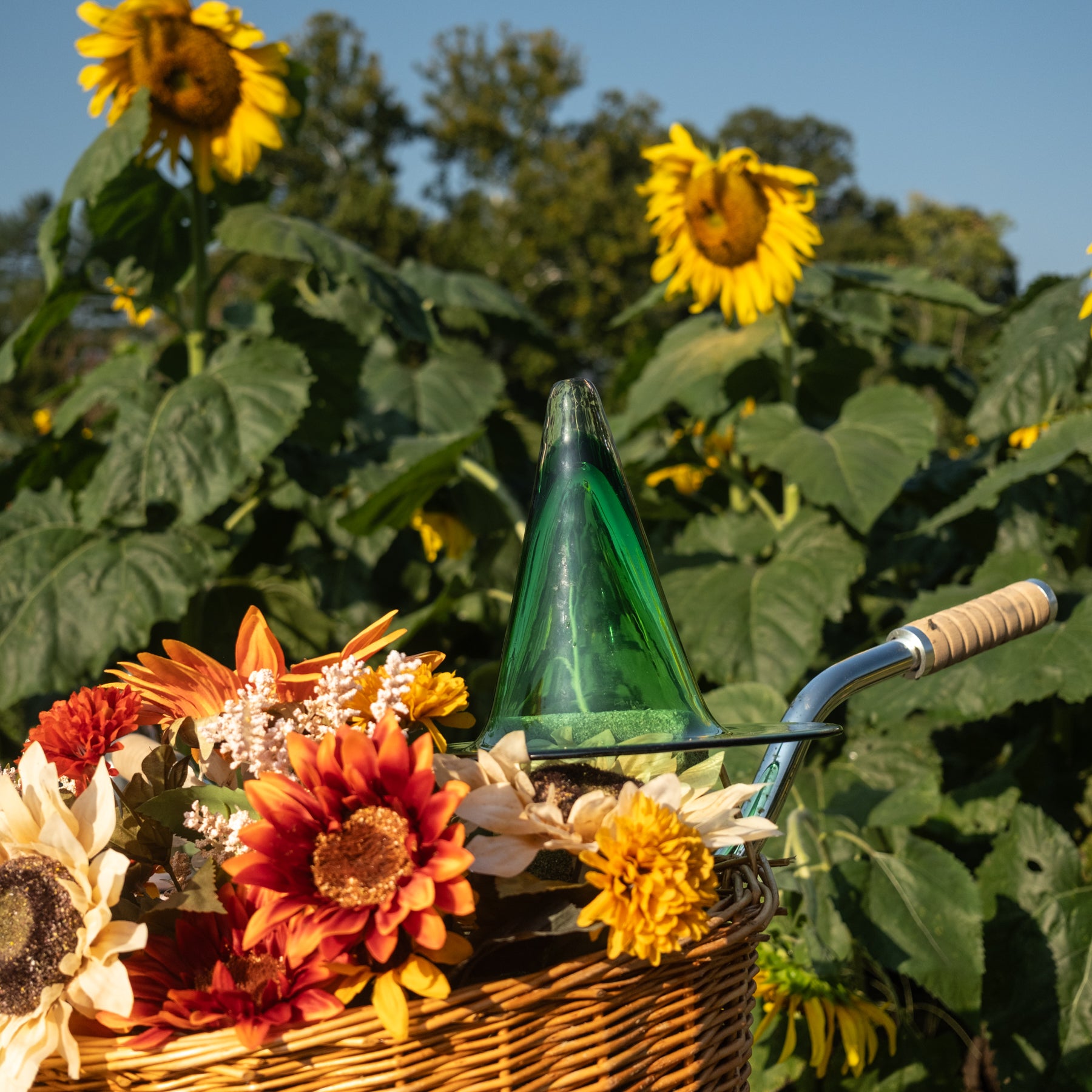 A green glass watering can with a curved metal handle sits in a wicker basket filled with sunflowers and autumn flowers, set against a sunny garden background with large sunflowers and green foliage.