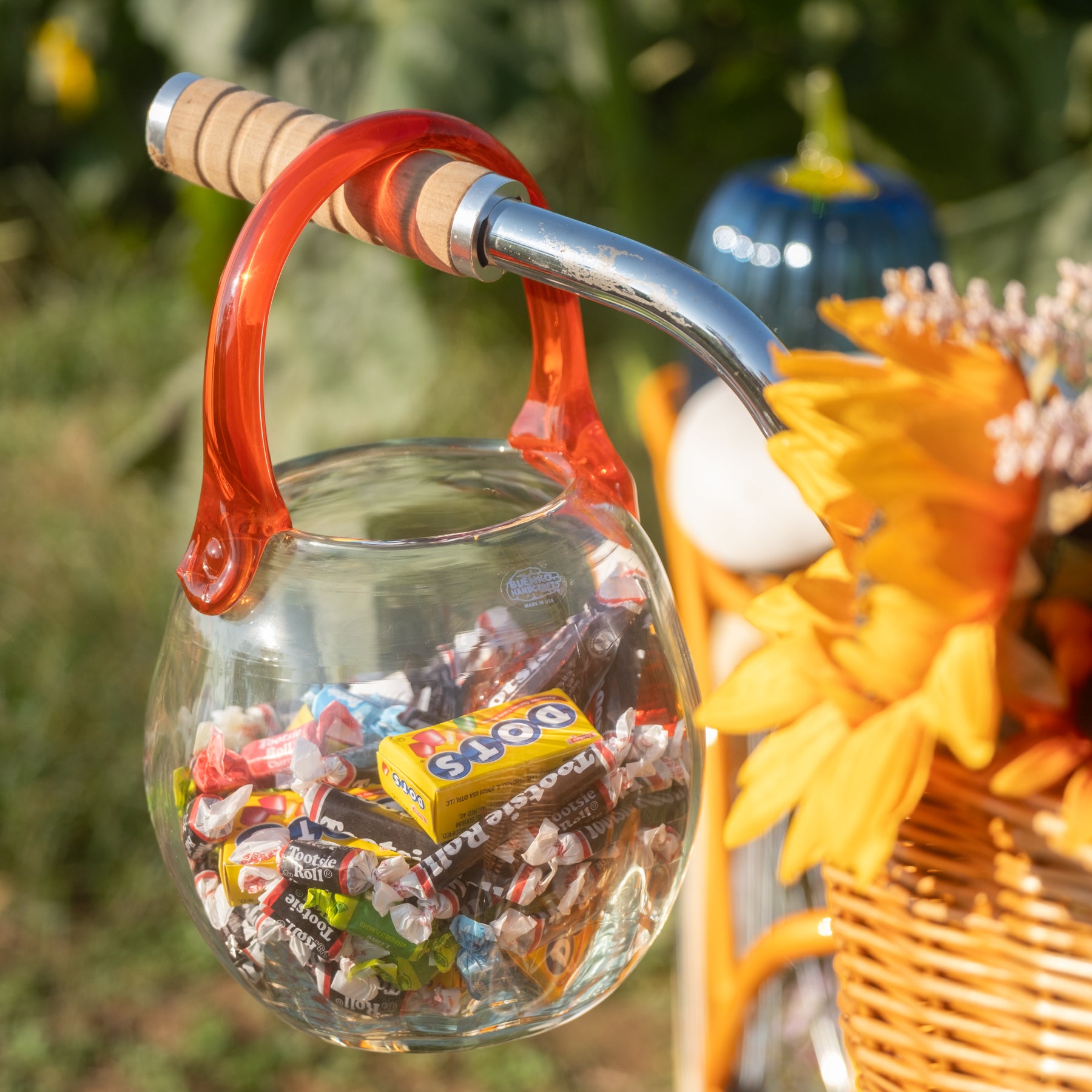 A clear glass bowl-shaped container with a translucent red handle and a light wood handle grip, hanging from a metal handlebar. The container is filled with assorted wrapped candies, including a visible Snickersa box, and is photographed outdoors with a blurred green background and a wicker basket of flowers at the right.