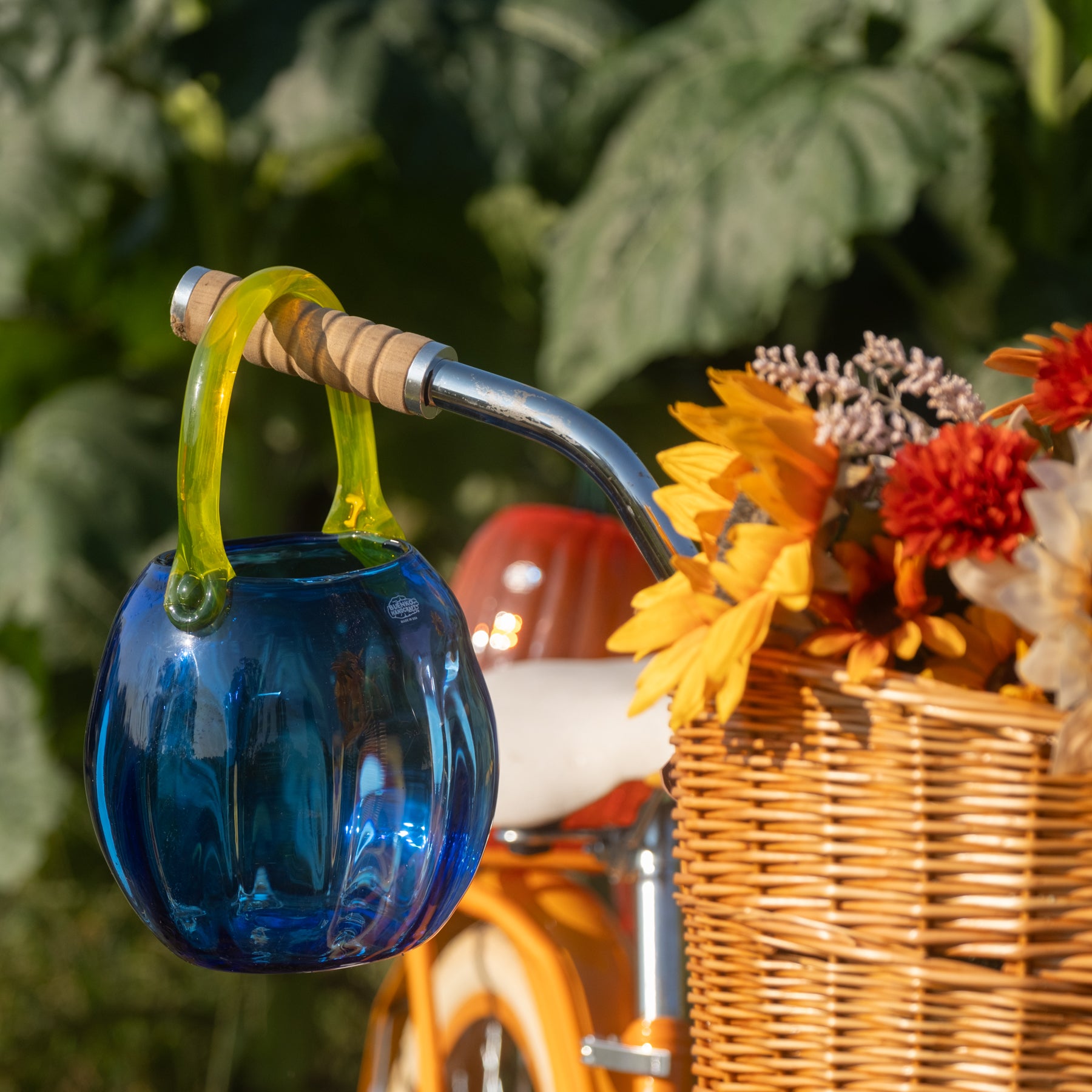 A blue ribbed glass hanging pot with a translucent green loop handle, attached to a wicker basket filled with orange and yellow flowers on a vintage-style bicycle outdoors.